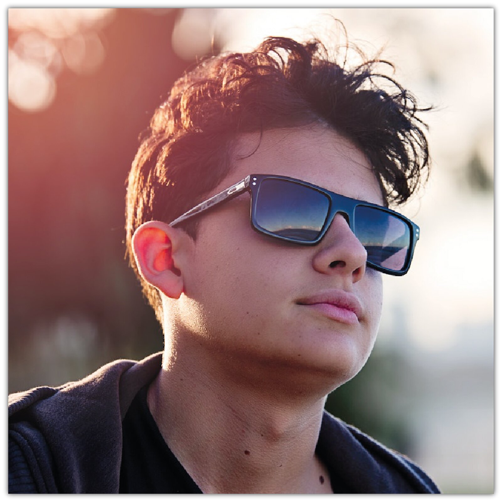 Zdjęcie przedstawia młodego chłopaka w bluzie i w okularach przeciwsłonecznych. Ma lekko uniesioną głowę i patrzy w dal. 