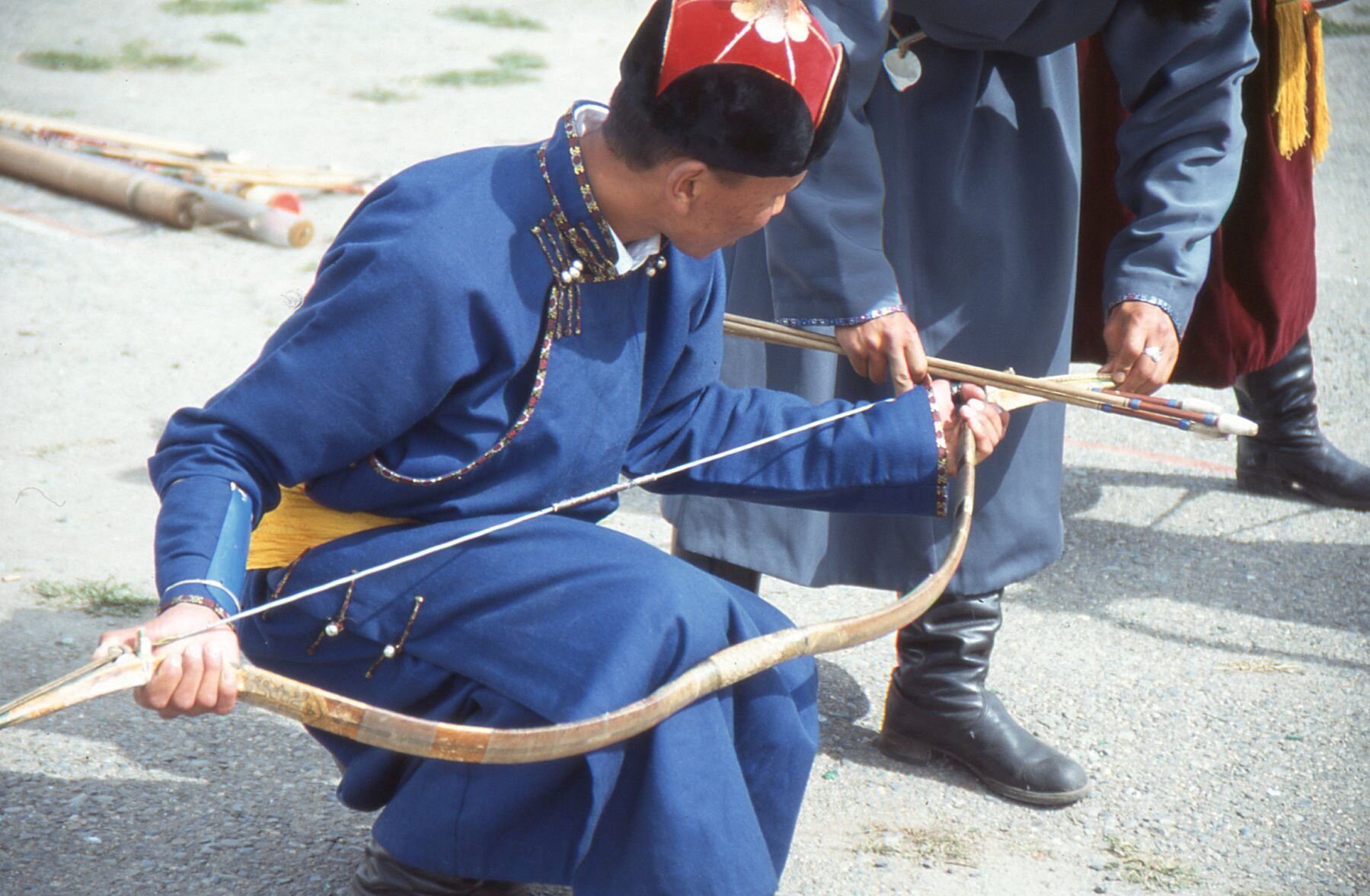 Łucznicy w tradycyjnych mongolskich strojach podczas dorocznego festiwalu sportowego Nadaam w Ułan Bator. W kadrze wyraźnie widoczny jest jeden, klęczący na jednym kolanie, trzymający oburącz łuk. Drugi widoczny jest od pasa w dół, jak podaje strzały pierwszemu. Noga trzeciego widoczna jest w górnym prawym rogu zdjęcia.