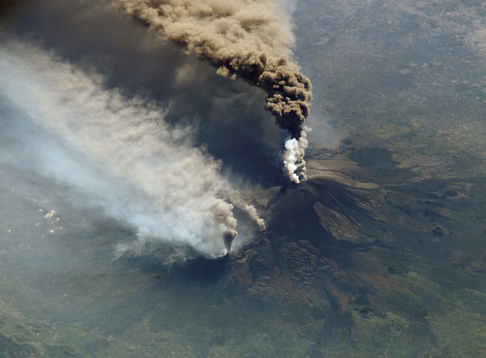 Fotografia wykonana z góry przedstawia wulkan podczas erupcji. Ze szczytu wulkanu wydobywa się chmura biało‑brazowych gazów i pyłów.