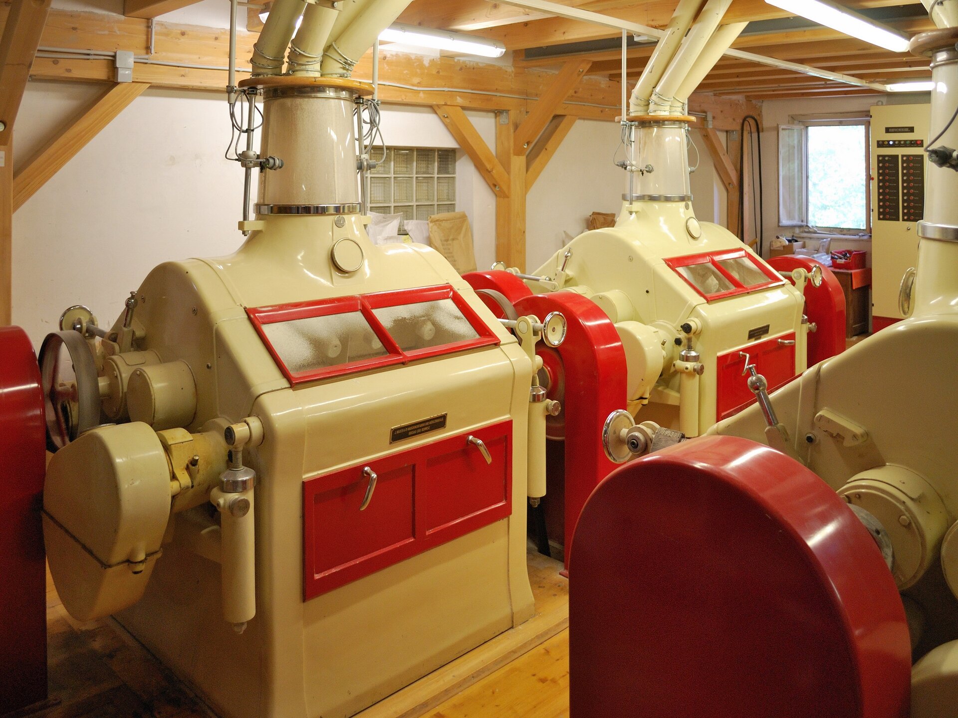 Zdjęcie przedstawia pomieszczenie z beżowymi mlewnikami walcowymi służącymi do rozdrabniania ziarna. Maszyny składają się z czterech ścian, na których widnieją czerwone, prostokątne drzwiczki z srebrnymi klamkami. Nad drzwiczkami znajduje się okienko w czerwonej ramie. Góra maszyny przypomina dwuspadziowy dach z kominem, z którego wychodzą rury pnące się do sufitu. Podłoga w pomieszczeniu jest drewniana. 