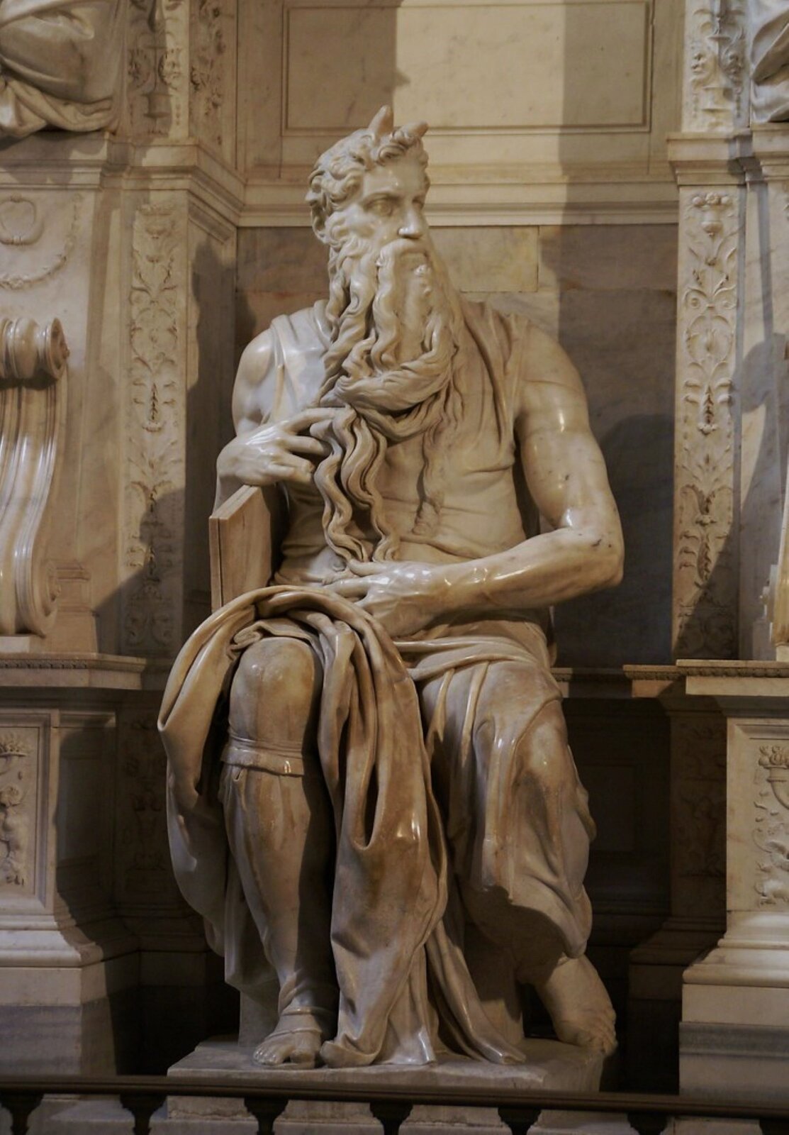 Ilustracja przedstawia rzeźbę siedzącego mężczyzny we wnęce pomiędzy filarami. Mocno udrapowana szata opada na nogi odsłaniając prawe kolano oraz ramiona. Mężczyzna jest budowy atletycznej i siedzi swobodnie, lewą rękę ma opartą o prawą nogę, a prawą rękę - o krawędź oparcia. Lewa noga jest nieco cofnięta do tyłu. Mężczyzna ma na głowie - zwróconej w lewą stronę - dwa niewielkie rogi .Głowę zwróconą w lewą stronę. Ma kręcone krótkie włosy i długą brodę.