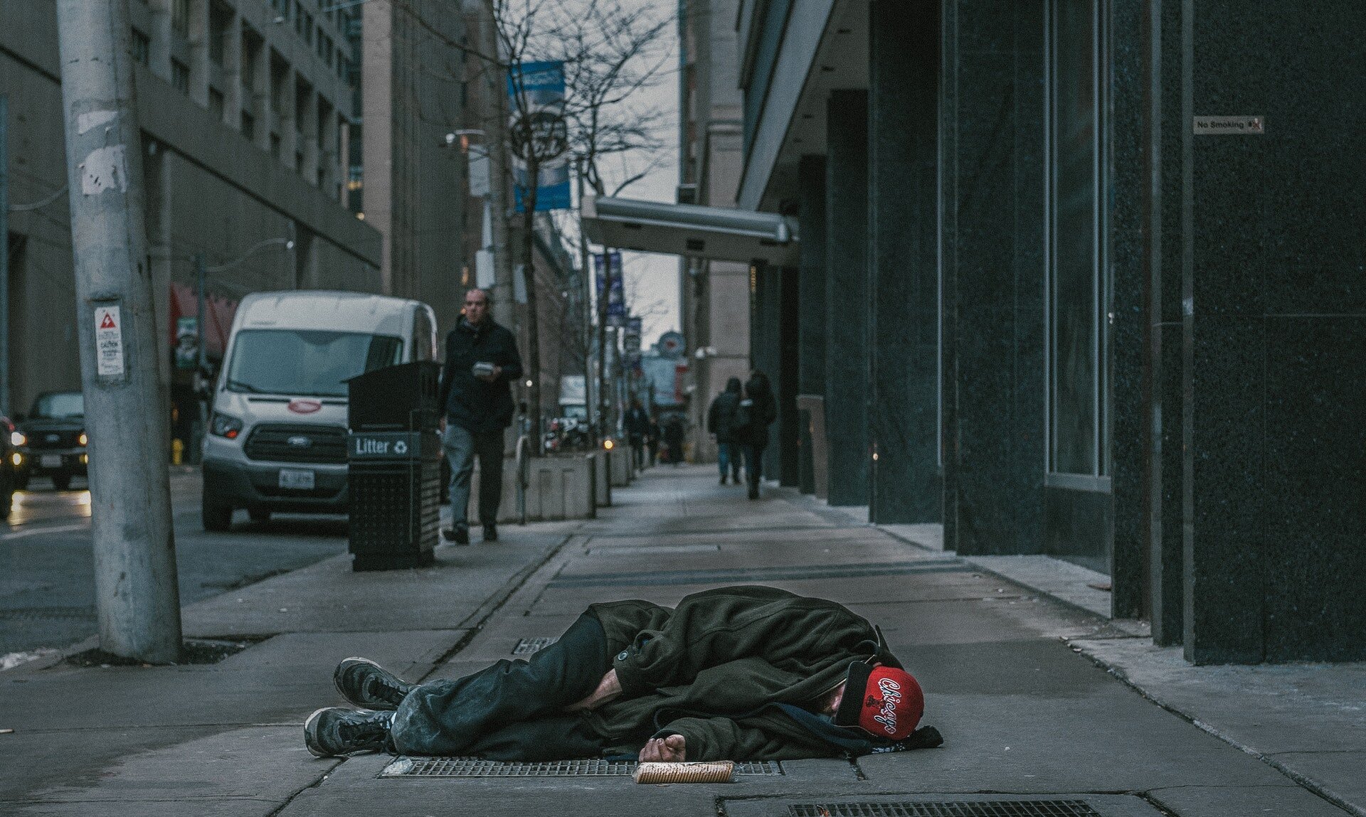 Zdjęcie przedstawia miejską ulicę. Na chodniku leży na lewym boku człowiek.