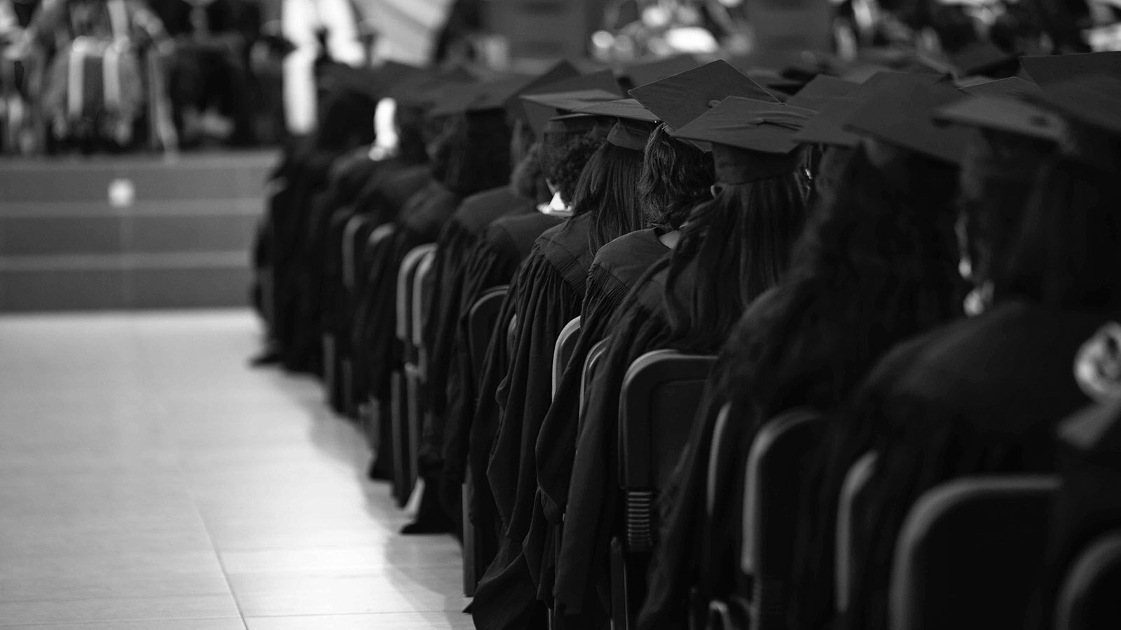 Na czarnobiałym zdjęciu uchwycono siedzących w ławkach studentów ostatniego roku ubranych w czarne togi i z czarnymi czapkami na głowach.