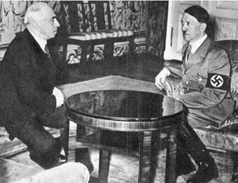 Zdjęcie przedstawia dwóch mężczyzn siedzących przy okrągłym stole. Po lewej, starszy, siwiejący w garniturze lekko pochylony do przodu. Na przeciwko niego siedzi mężczyzna w średnim wieku, w mundurze ze swastyką na ramieniu, ma krótki, charakterystyczny wąs pod nosem i przygładzone, zaczesane na bok włosy.