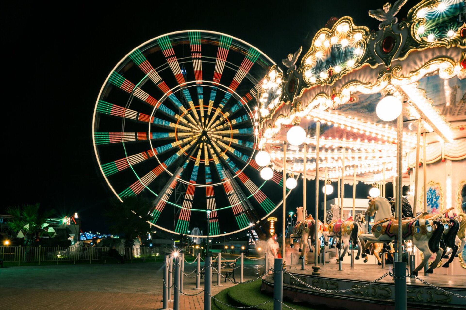 Ilustracja ukazuje park rozrywki oświetlony wieczornymi światłami. Na pierwszym planie widoczna jest karuzela z kolorowymi konikami.