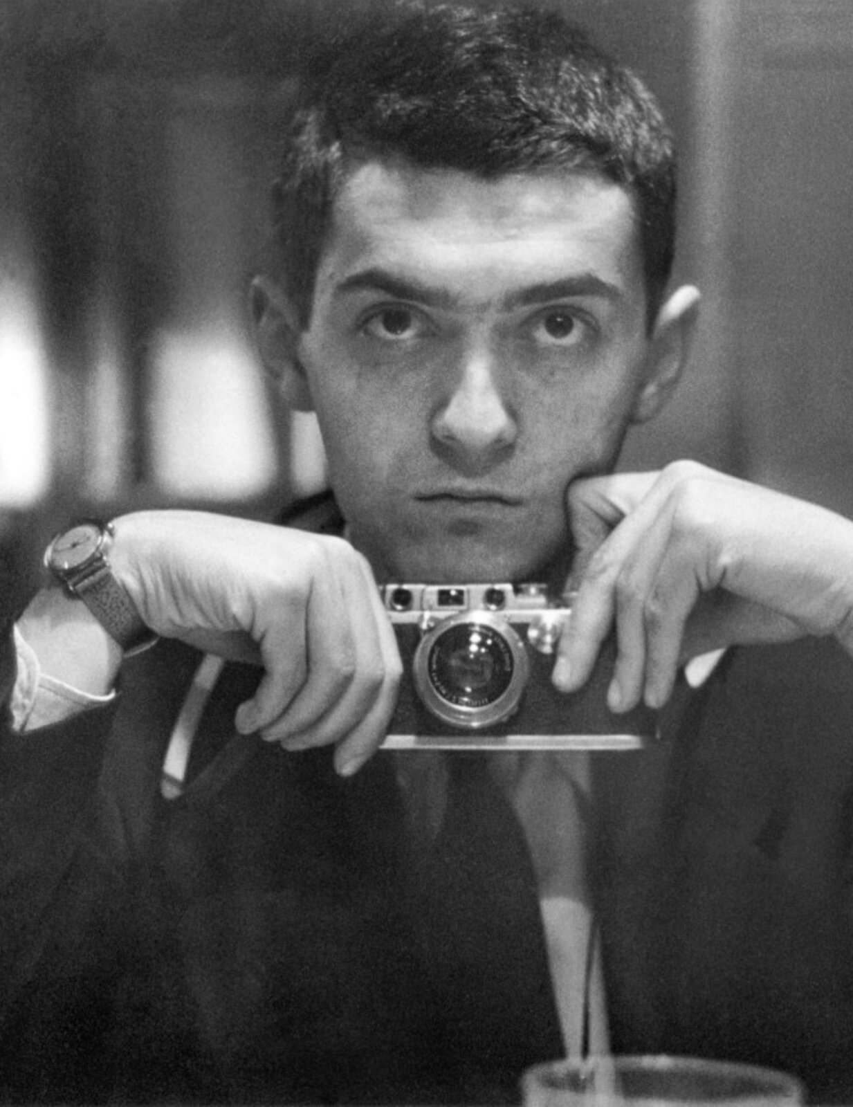 Sfotografowana osoba to młody mężczyzna o krótko ściętych włosach, który patrzy prosto w obiektyw. Jego oczy są szeroko otwarte, na jego czole widoczne są poprzeczne zmarszczki, a wąskie usta są ściągnięte. W dłoniach, pod podbródkiem, trzyma aparat fotograficzny, którym zrobił sobie zdjęcie w lustrze. Mężczyzna ma na sobie marynarkę, koszulę oraz krawat. Tło jest rozmyte.