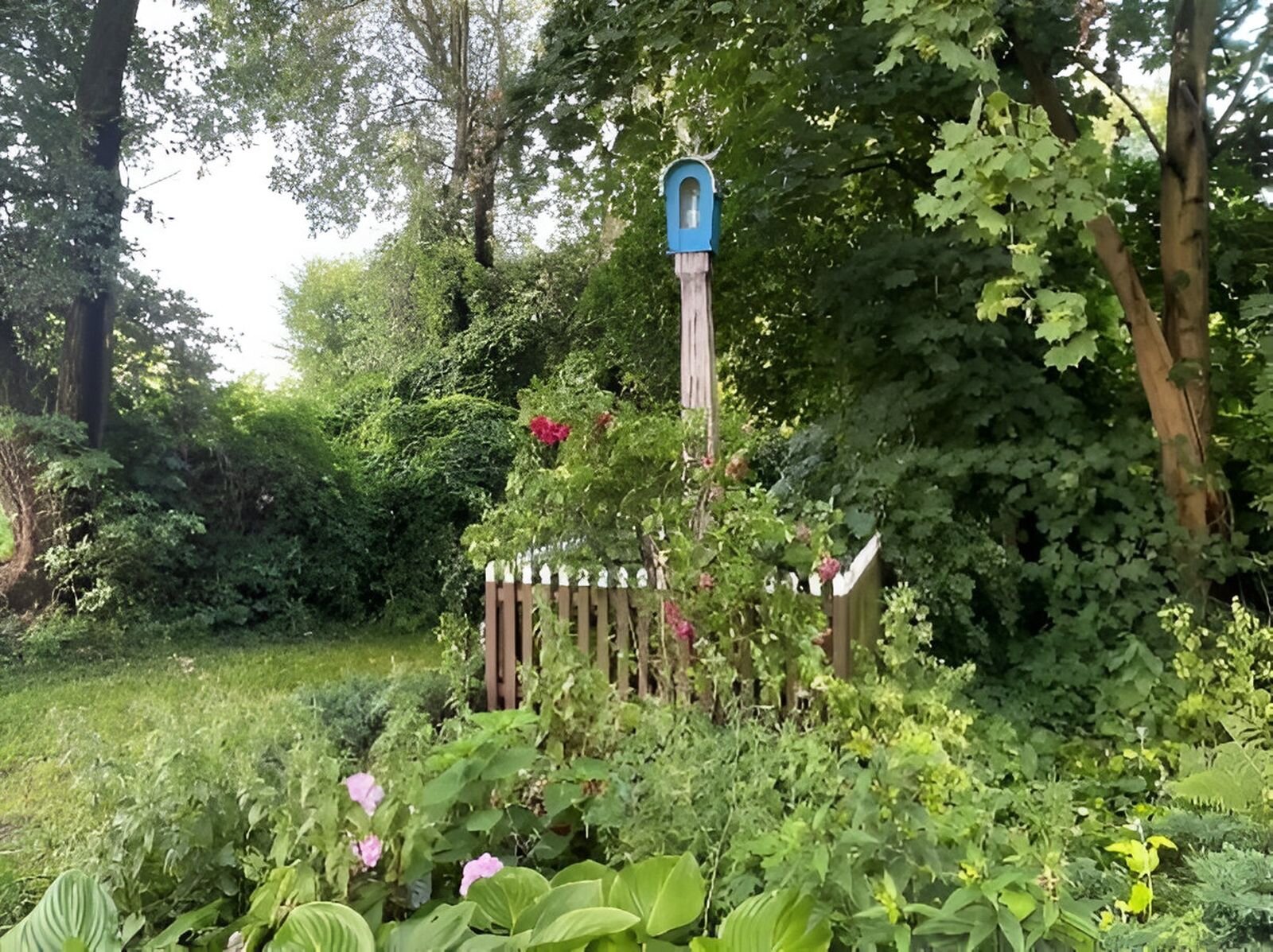 Fotografia przedstawia kapliczkę. Wokół niej zarośla i drewniany płotek, wokół którego są kępiaste kwiaty. Za kapliczką drzewa.