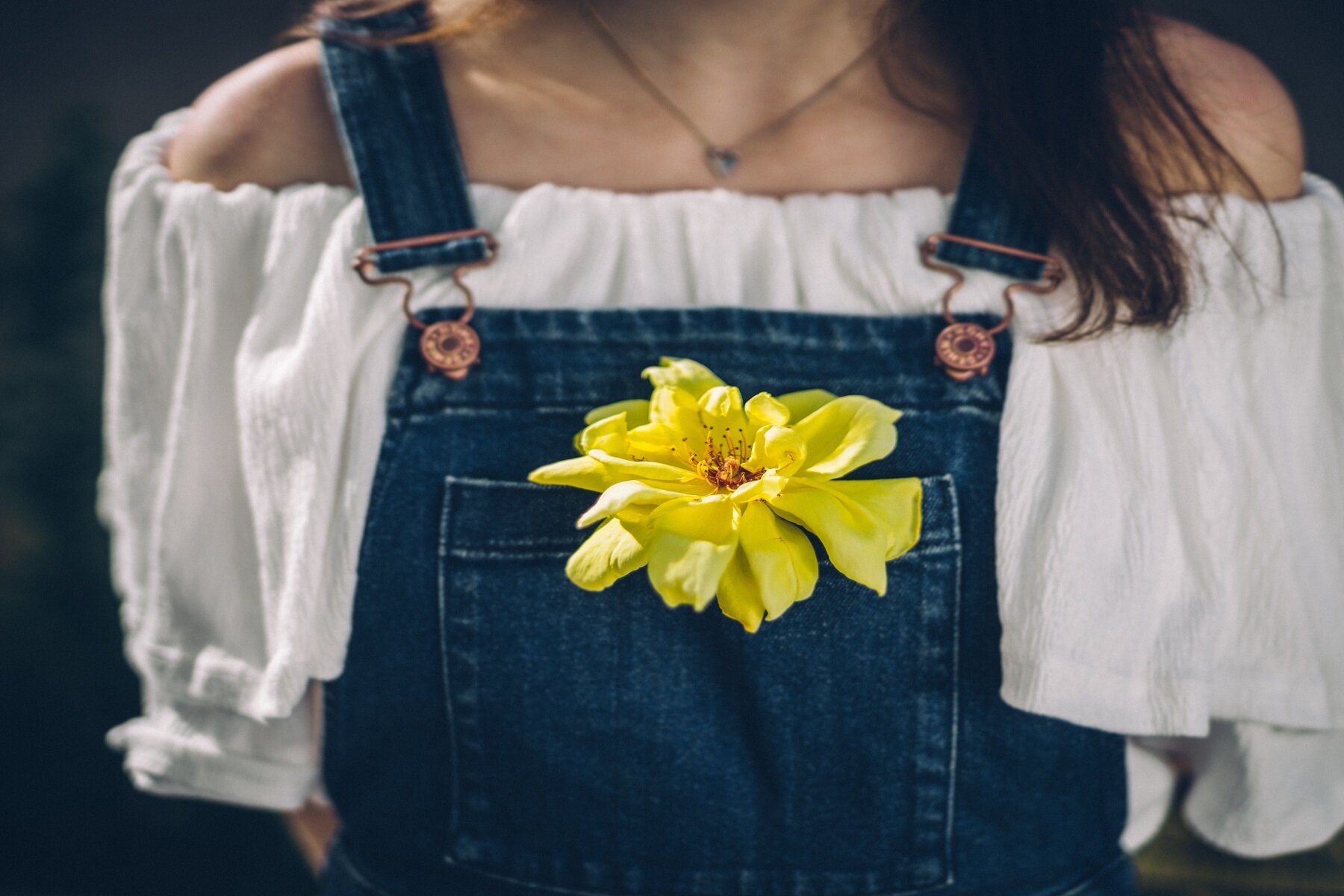 Zdjęcie przedstawia fragment dziewczyny od pasa do szyi. Dziewczyna ubrana jest w jeansowe ogrodniczki i białą koszulę z odkrytymi ramionami. Na szyi ma zawieszony łańcuszek. W kieszonce ogrodniczej, na piersi, widoczny jest żółty, lekko zwiędnięty kwiatek. 