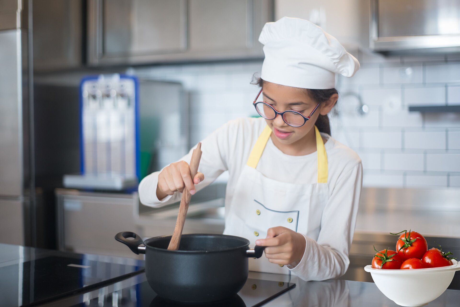 Zdjęcie przedstawia dziewczynkę stojącą przy blacie roboczym z płytą indukcyjną, na której stoi czarny garnek. Dziewczynka ma na sobie fartuszek oraz nakrycie głowy kucharza. Długie ciemne włosy ma upięte z tyłu. Dziewczynka ma lekko ugięte ręce. W prawej trzyma drewnianą łyżkę zanurzoną w garnku, zaś lewą trzyma uchwyt garnka. Na prawo od niej, na blacie, stoi miska z pomidorami. W tle widoczne rozmyte meble kuchenne.