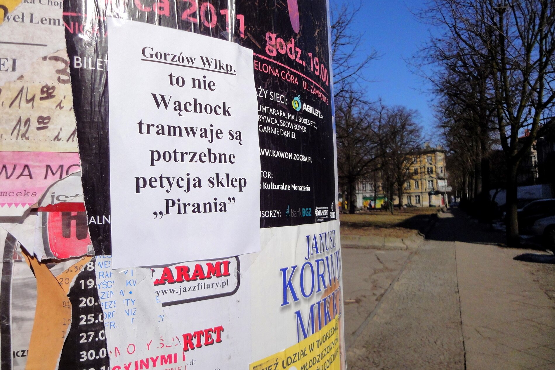 Zdjęcie przedstawia fragment słupa ogłoszeniowego stojącego na ulicy. Zawieszona jest na niej kartka z napisem: Gorzów Wielkopolski to nie Wąchock tramwaje są potrzebne petycja sklep Pirania.