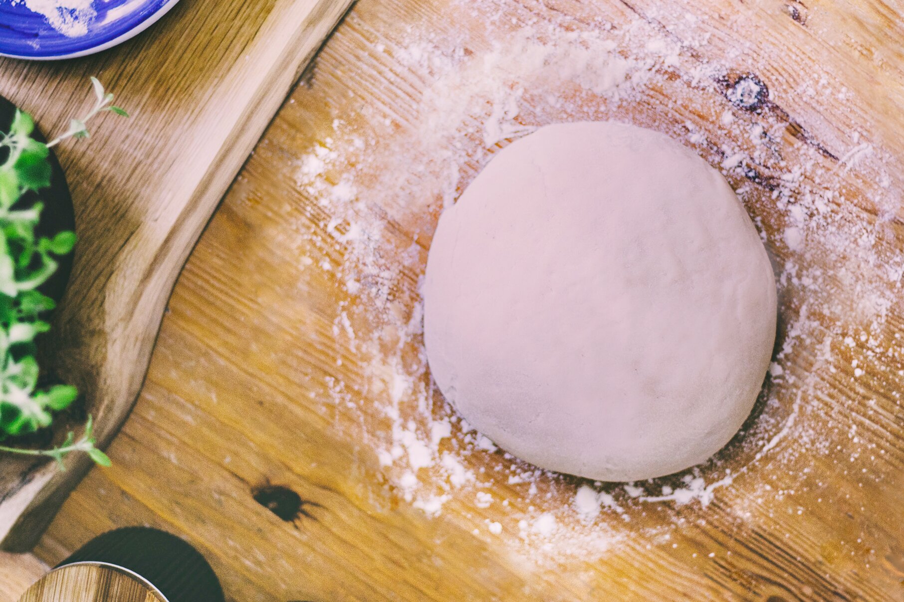 Zdjęcie przedstawia krok pierwszy formowania precli. Ujęcie wykonane jest od góry. Na pierwszym planie widoczna jest jasna kula ciasta obsypana mąką. Leży ona na drewnianym ciemnobrązowym blacie. Dookoła ciasta rozsypana jest biała mąka. W lewym dolnym rogu zdjęcia dostrzec można fragment czarnej puszki z drewnianym wieczkiem. W lewej części grafiki widoczny jest zarys czarnej doniczki z zieloną rośliną, a obok znajduje się fragment niebieskiego talerza. Wszystkie te przedmioty leżą na drewnianej jasnobrązowej desce.