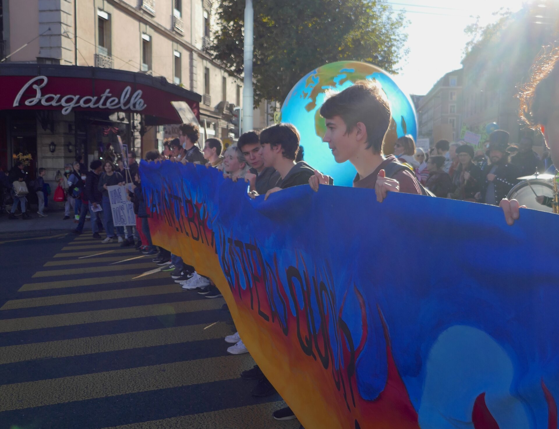 Zdjęcie przedstawia młodzież uczestniczącą w proteście. Młodzież trzyma w rękach duży plakat, który jest kolorowy - od żółtego na dole, przechodząc przez czerwony na górze, z niebieskim obszarem i czarnym napisem. W tle widoczny jest duży globus oraz nazwa sklepu "Bagatelle".