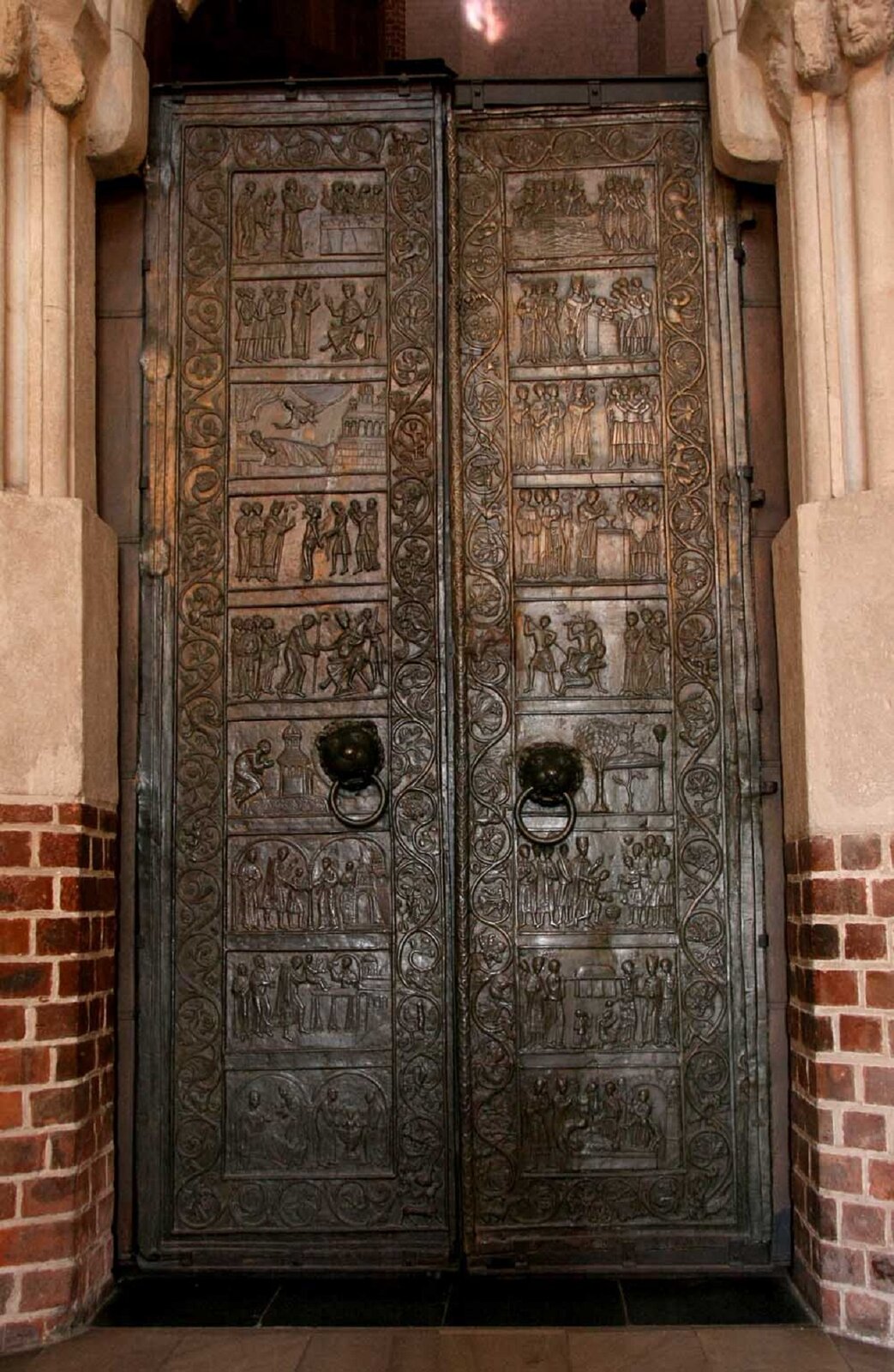 Zdjęcie przedstawia stare drzwi wykonane z brązu. Na drzwiach widnieje wytłoczona historyjka obrazkowa z udziałem świętych.