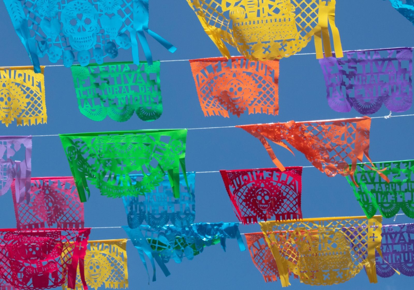 Zdjęcie przestawia ażurowe chorągiewki - wycinanki zawieszone na sznurkach jedna obok drugiej. Sznurki z chorągiewkami wypełniają cały kadr, są ułożone poziomo. Na chorągiewkach wycięte są różne wzory  i napisy, niektóre mają powiewające na wietrze frędzelki. Jest sześć rzędów sznurków z chorągiewkami, na każdym sznurku powiewają 2‑4 wycinanki. Tło jest niebieskie.