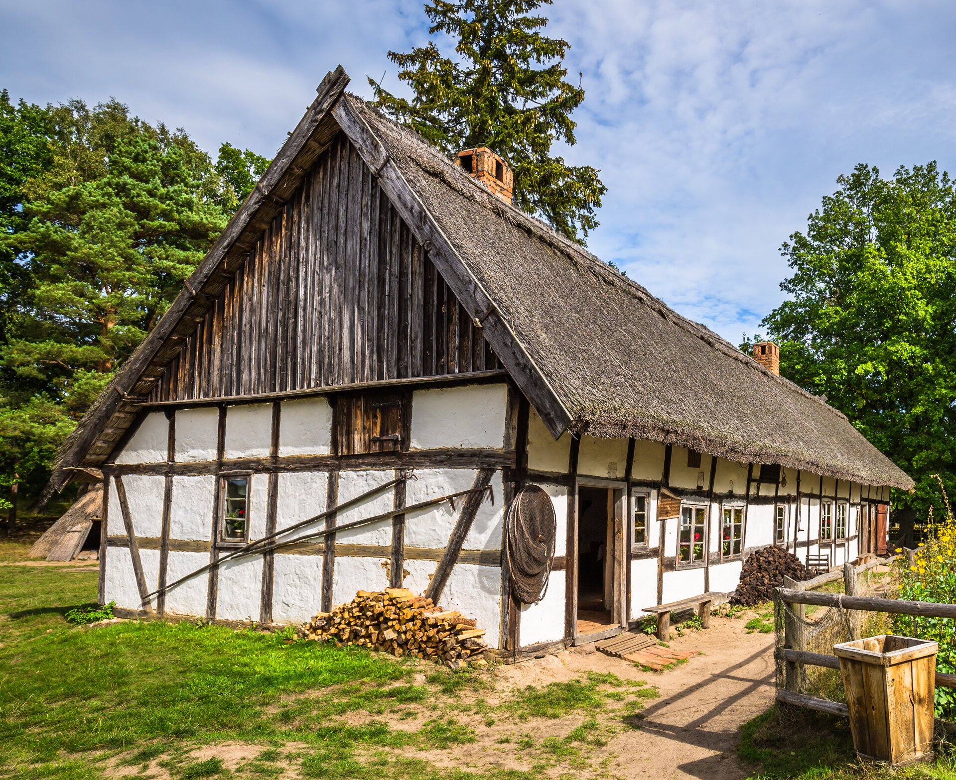 Kliknij, aby powiększyć. Zdjęcie przedstawia chatę. Jest jednopiętrowa i posiada dwuspadowy dach pokryty trzciną. Budynek ma konstrukcję szkieletową złożoną z drewnianych belek wypełnionych murem, który jest bielony. Wokół domu znajduje się trawnik, z prawej strony zdjęcia jest płot.
