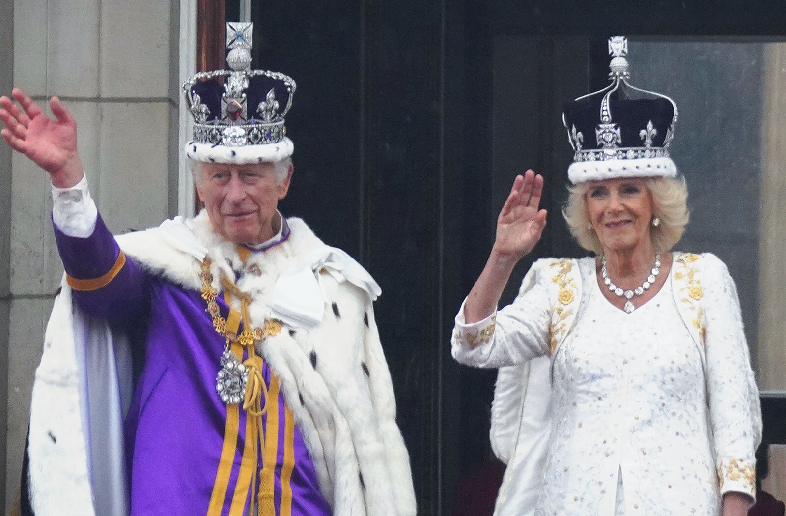 Zdjęcie przedstawia króla Wielkiej Brytanii Karola III oraz królową Kamillę. Po lewej stronie widoczny jest stojący mężczyzna w starszym wieku, z krótkimi, siwymi włosami, ubrany w fioletową szatę zdobioną złotymi pasami i pelerynę z białego futra. Na głowie ma królewską koronę wysadzaną kamieniami szlachetnymi, a na piesi medalion na złotym łańcuchu. Prawą rękę ma uniesioną do góry w geście pozdrowienia. Na prawo od niego widoczna jest stojąca kobieta w starszym wieku, z półdługimi siwymi włosami, wywiniętymi na końcach, ubrana w białą suknię z białą peleryną wysadzaną kamieniami szlachetnymi. Na głowie ma królewską koronę wysadzaną kamieniami szlachetnymi, a na szyi kolię z kamieni szlachetnych. Prawą rękę ma uniesioną do góry w geście pozdrowienia, a lewą opuszczoną wzdłuż ciała.
