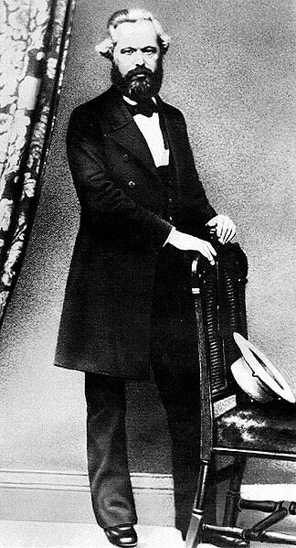 Czarno‑biała fotografia portretowa mężczyzny w średnim wieku, obejmuje całą sylwetkę. Ma na sobie  pantofle, ciemne spodnie garniturowe, kamizelkę, białą koszulę i długą marynarkę lub płaszcz. Mężczyzna ma gładkie zaczesane do tyłu włosy i bujną brodę, pozuje opierając ręce o oparcie krzesła, na którego siedzeniu leży kapelusz.