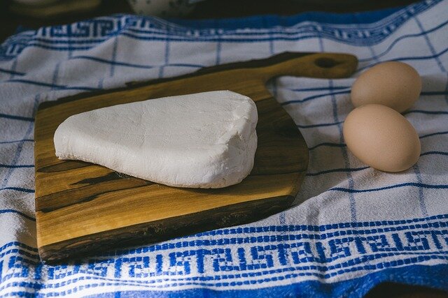 Zdjęcie przedstawia biały ser w sercowatej formie, ułożony na drewnianej desce. Na prawo widoczne dwa kurze jaja. Całość ułożona na białej ściereczce z motywem meandra i kratką w kolorze niebieskim.