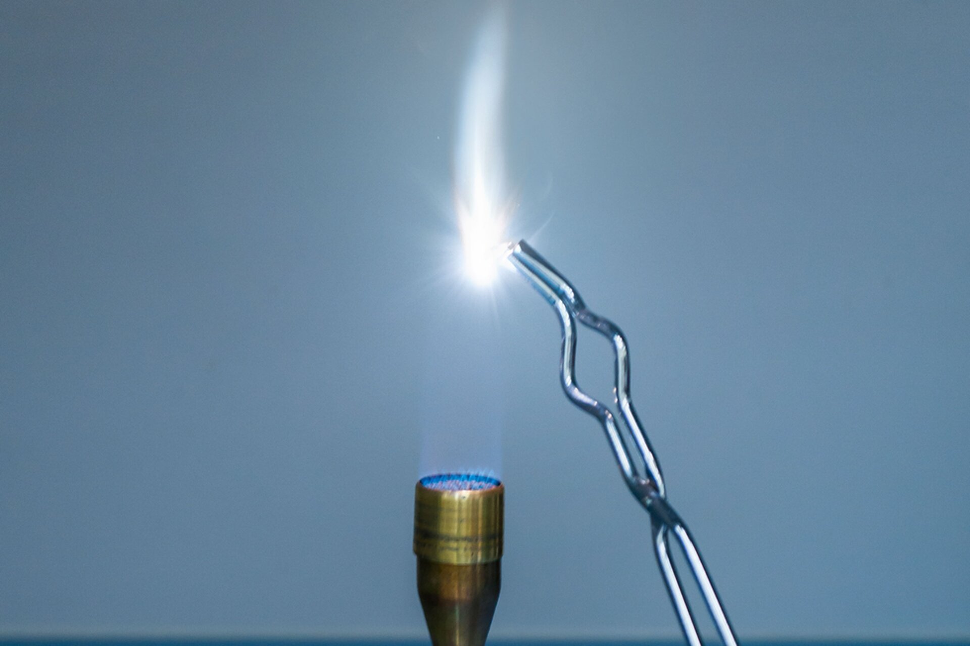Zdjęcie przedstawia palnik gazowy, nad którym ręka eksperymentatora trzyma długie szczypce, których koniec znajduje się w zasięgu płomienia. Trzymana w szczypcach wstążka magnezowa spala się białym oślepiającym płomieniem.