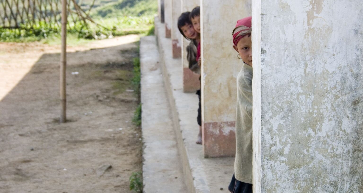 Fotografia przedstawia zajmujący jej prawą połowę fragment murowanej budowli ukazanej w perspektywie. Składa się na nią podwyższony o jeden stopień podest i sześć równoległych ścianek ustawionych na nim jedna za drugą. Są one pomalowane w dolnej części ciemnoróżową, a powyżej białą farbą, która odpada. Z lewej strony fotografia przedstawia podwórko z wkopanym na środku bambusowym drągiem i wyraźnym cieniem budynku. Zamknięte jest ono częściowo z tyłu drewnianym płotem ukazanym w słońcu. Za pierwszą ścianką ukryte jest azjatyckie dziecko tak, że widać tylko prawą połowę jego ciała. Ubrane jest ono w jasny sweter, czerwoną chustkę skrywającą włosy i nosi w uchu kolczyk w kształcie kółka. Przygląda się ono uważnie widzowi jednym okiem. W głębi zza drugiej ścianki wygląda ciekawie jeszcze dwoje uśmiechniętych dzieci.