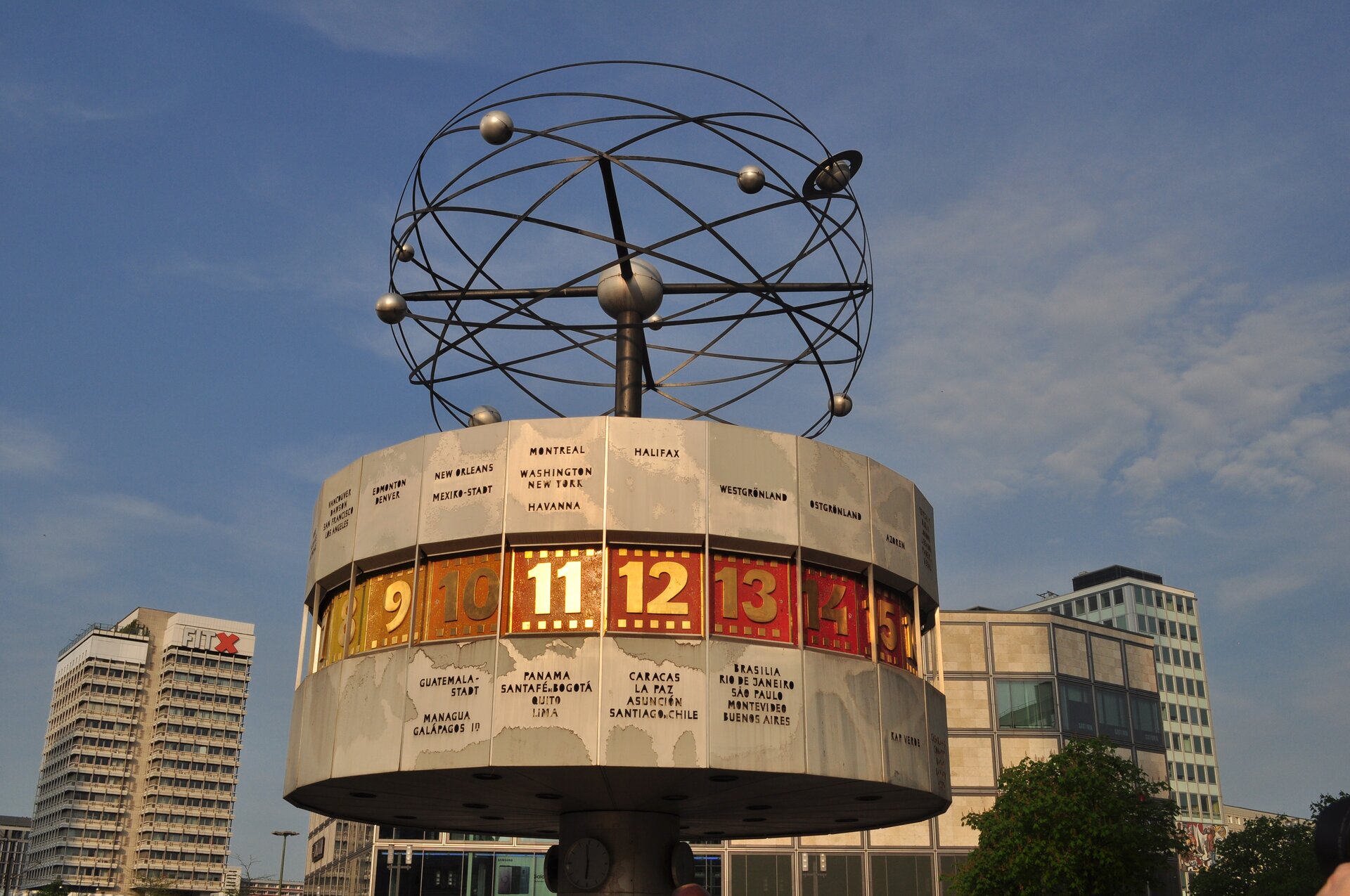 Ilustracja przedstawia pomnik. To Zegar Czasu na Świecie w Berlinie. Na owalnej metalowej podstawie widoczne są liczby. Na niej widoczna jest konstrukcja na planie kuli przedstawiająca ruch planet po osiach. Poniżej ilustracji znajduje się nagranie oraz transkrypcja.