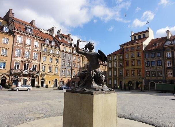 Zdjęcie przedstawia pomnik syreny, czyli kobiety która zamiast nóg ma rybi ogon. W lewej dłoni trzyma tarczę. Prawa ręka uniesiona nad głową, trzyma w niej miecz.  Stoi na cokole. Za nią jest brukowany plac i rzędy kamienic. 