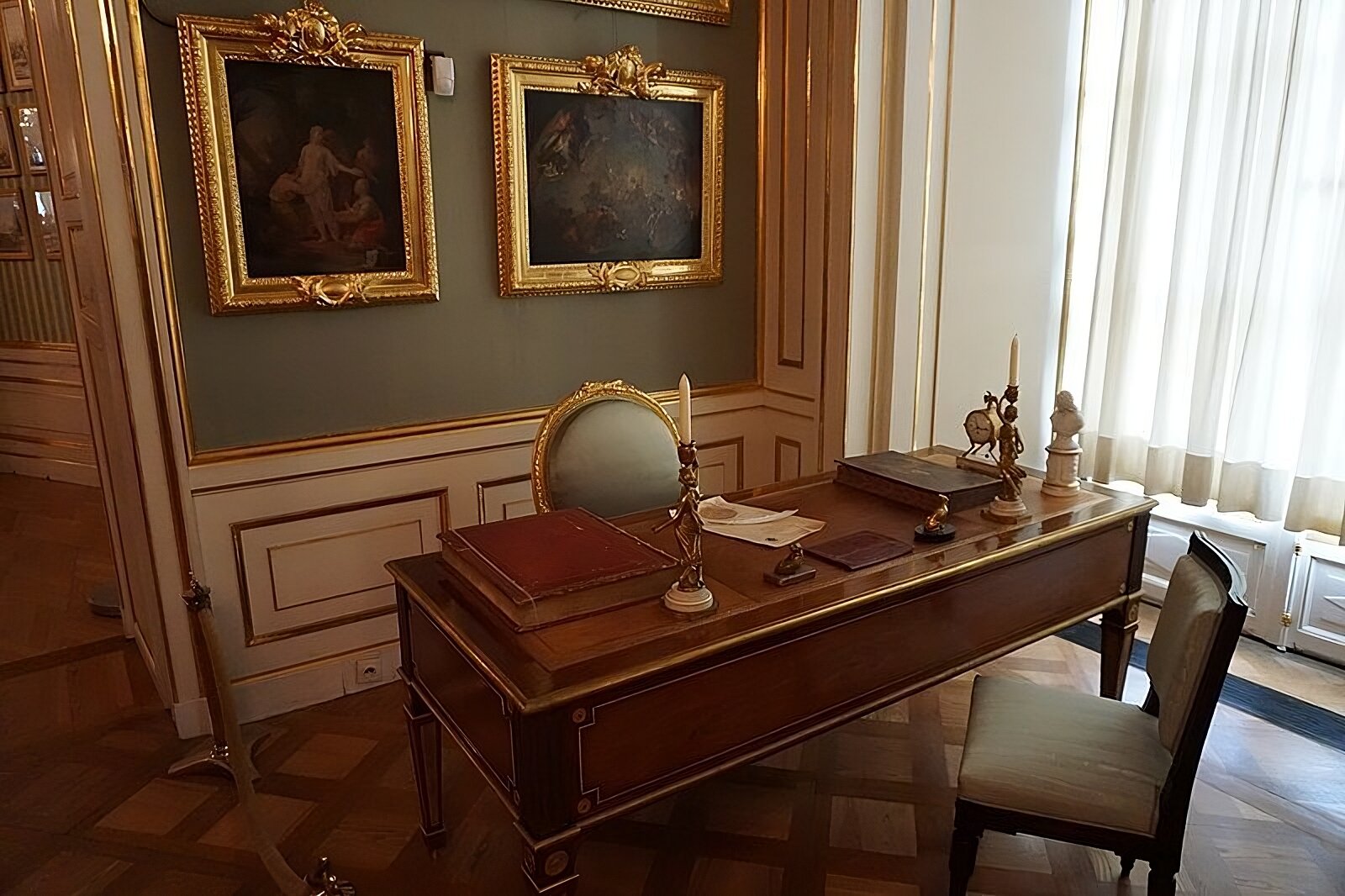 Zdjęcie przedstawia w centrum fotografii biurko króla Stanisława Augusta Poniatowskiego. Znajdują się na nim dwie księgi, dokument z pieczęcią królewską z położonym na nim gęsim piórem, dwa przyciski do papieru z figurkami psów, teczka na bibułki osuszające atrament, dwa mosiężne kandelabry, mosiężny zegar oraz figurka Voltaire’a [czyt.: Woltera]. Na ścianie za biurkiem dwa obrazy w złoconych ramach. Pod ścianą za biurkiem krzesło ze złotym oparciem obite szarą tkaniną, z drugiej strony biurka ciemne krzesło obite jasną tkaniną.