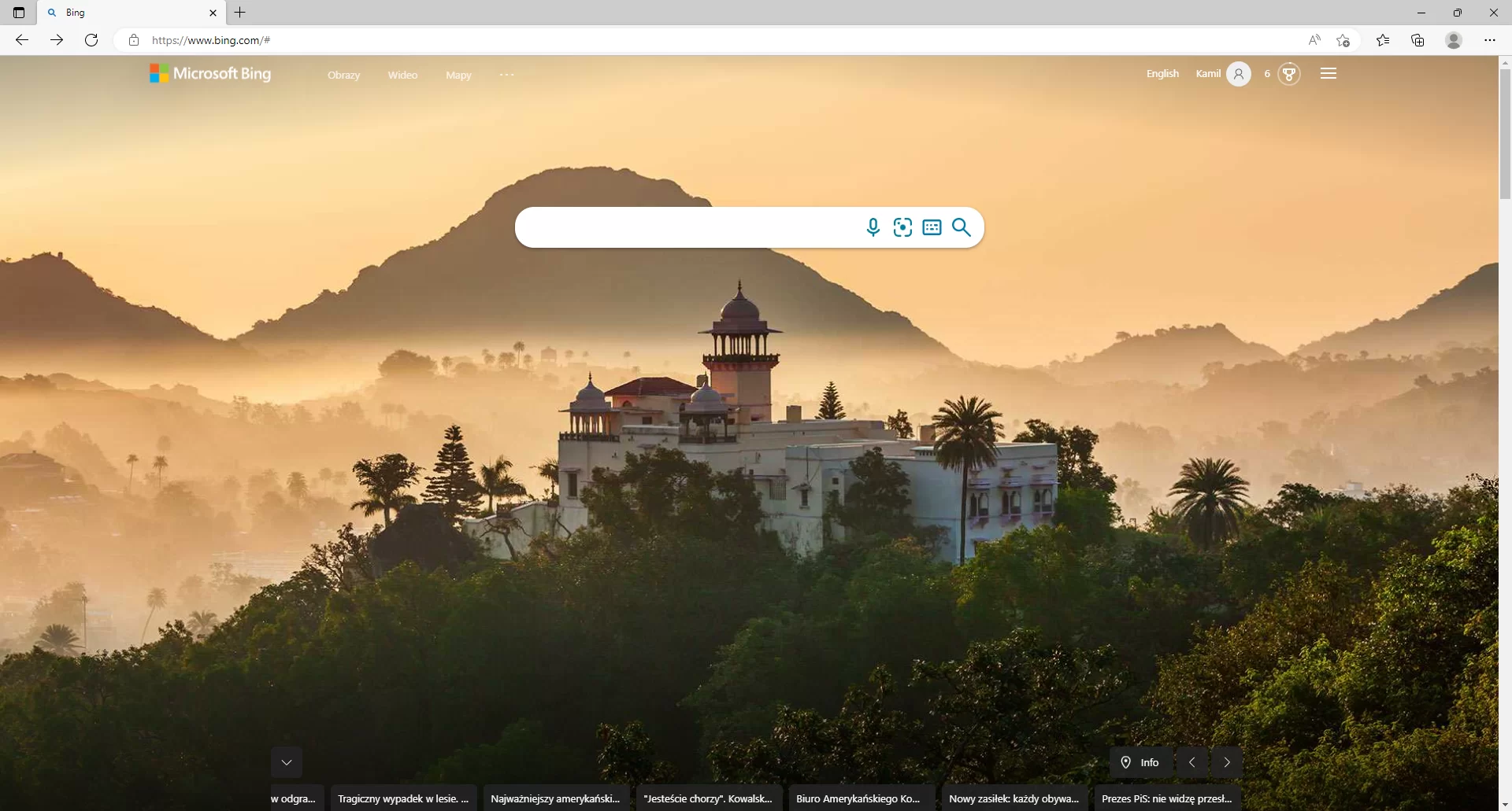 Zrzut okna przeglądarki  strony startowej Microsoft Edge. U góry widoczna jest jedna otwarta karta Bing. Poniżej znajduje się pasek adresowy z adresem "https://www.bing.com/#". W głównej części u góry znajduje się logo czterokolorowej flagi z podpisem Microsoft Bing oraz zakładki "Obrazy", "Wideo", "Mapy", zmiana języka "English", "Kamil", trofea oraz menu. Na środku znajduje się pasek wyszukiwania. Jako tapeta ustawiony jest orientalny budynek na wzgórzu wśród drzew. Są tam palmy, drzewa iglaste oraz liściaste. Budynek posiada trzy wieże - jedną wysoką i dwie niskie. W większości jego dachy są płaskie. Za nim rozpościera się widok na zamglone wzgórza. U dołu strony znajdują się wiadomości z częściowo widocznymi nagłówkami "Tragiczny wypadek w lesie", "Najważniejszy amerykański..", "Jesteście chorzy". Kowalsk...", "Biuro Amerykańskiego Ko...", "Nowy zasiłek: każdy obywa...", "Prezes PiS: nie widzę przesł...".
