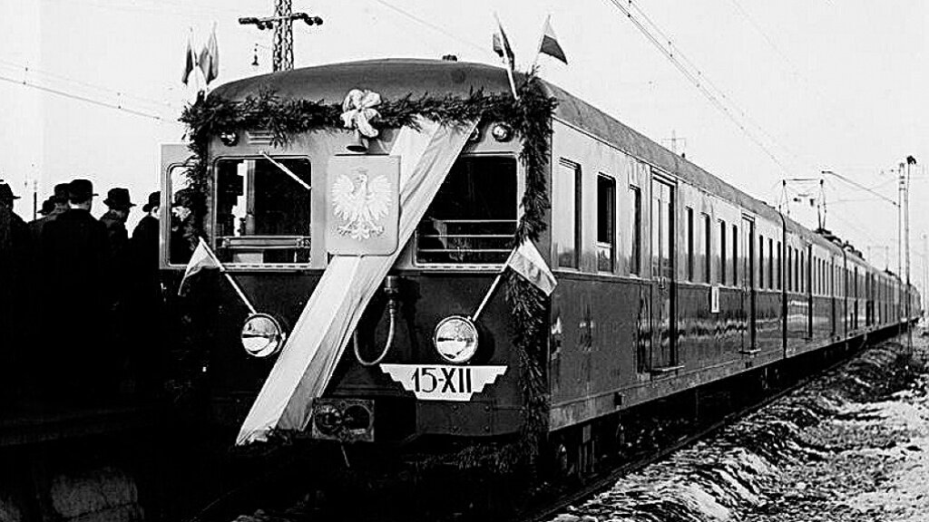 Zdjęcie przedstawia pociąg. Po lewej stronie są sylwetki osób w kapeluszach. Przód pociągu jest ozdobiony między innymi polskim godłem oraz wstęgą w kolorach narodowych. 