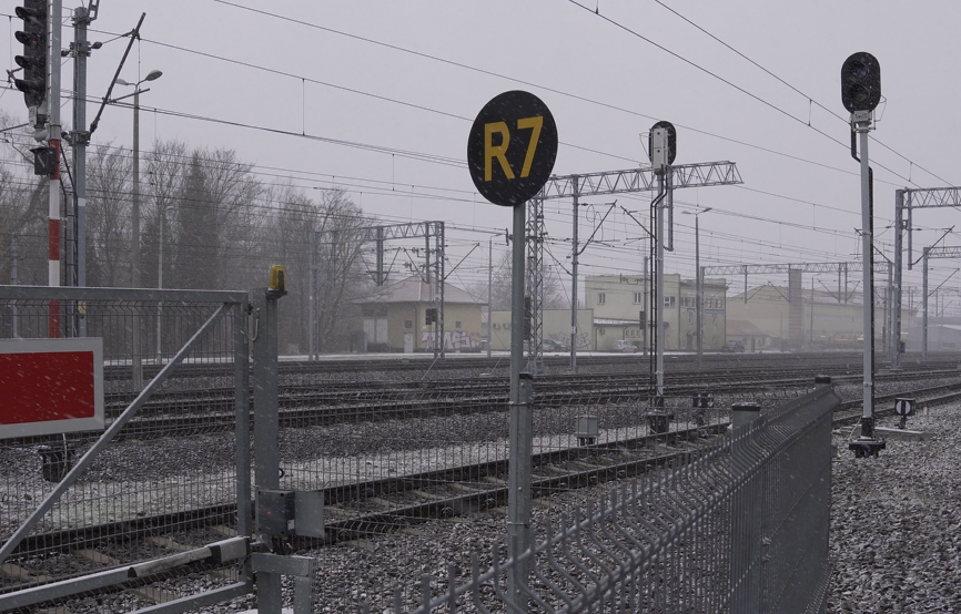 Zdjęcie przedstawia znak stojący przy torach kolejowych. Na metalowym słupku znajduje się okrągła czarna tarcza z żółtym napisem eR siedem.