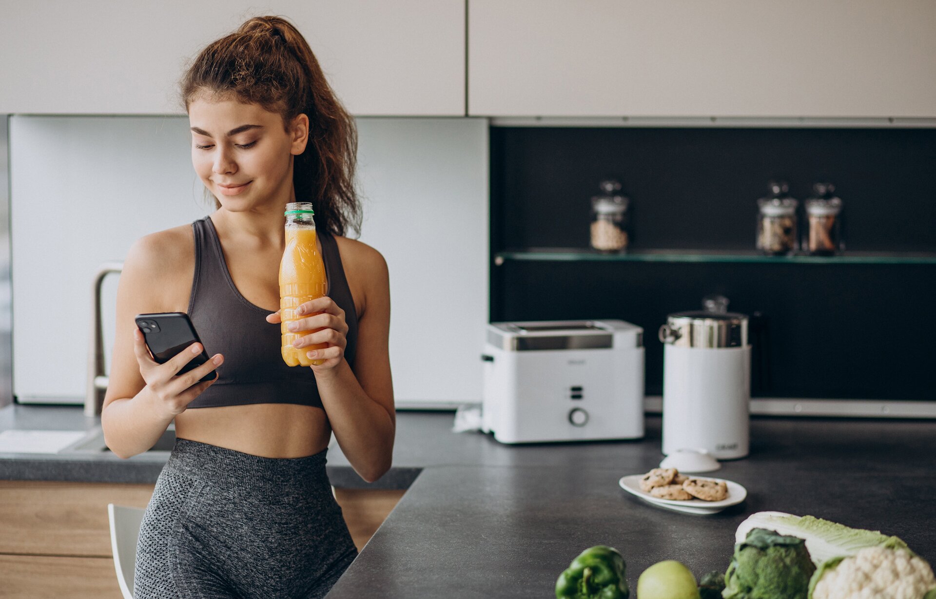 Zdjęcie przedstawia kobietę w młodym wieku w stroju sportowym, która stoi przy blacie kuchennym. W dłoni trzyma telefon, w który patrzy oraz butelkę z sokiem w kolorze pomarańczowym.