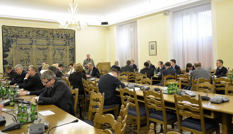 Zdjęcie przedstawia dużą salę. Są tu ustawione w trzech rzędach stoły, przy których siedzą posłowie i posłanki. Na końcu sali znajduje się stół prezydialny i mównica, przy której stoi mężczyzna.