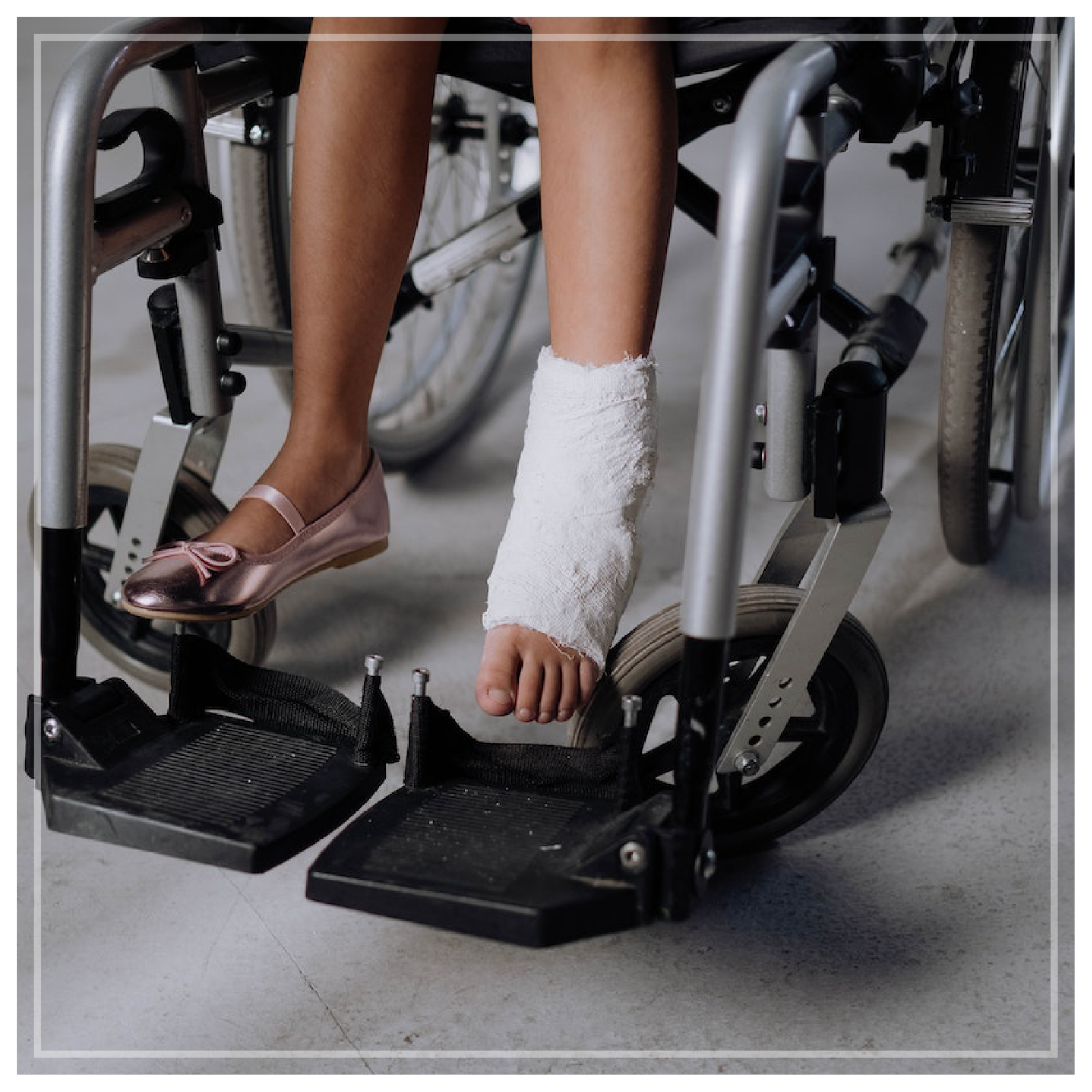 Zdjęcie nóg dziewczynki siedzącej na wózku inwalidzkim. Dziewczynka na jednej nodze ma różową balerinkę, a na drugiej ma założony gips.