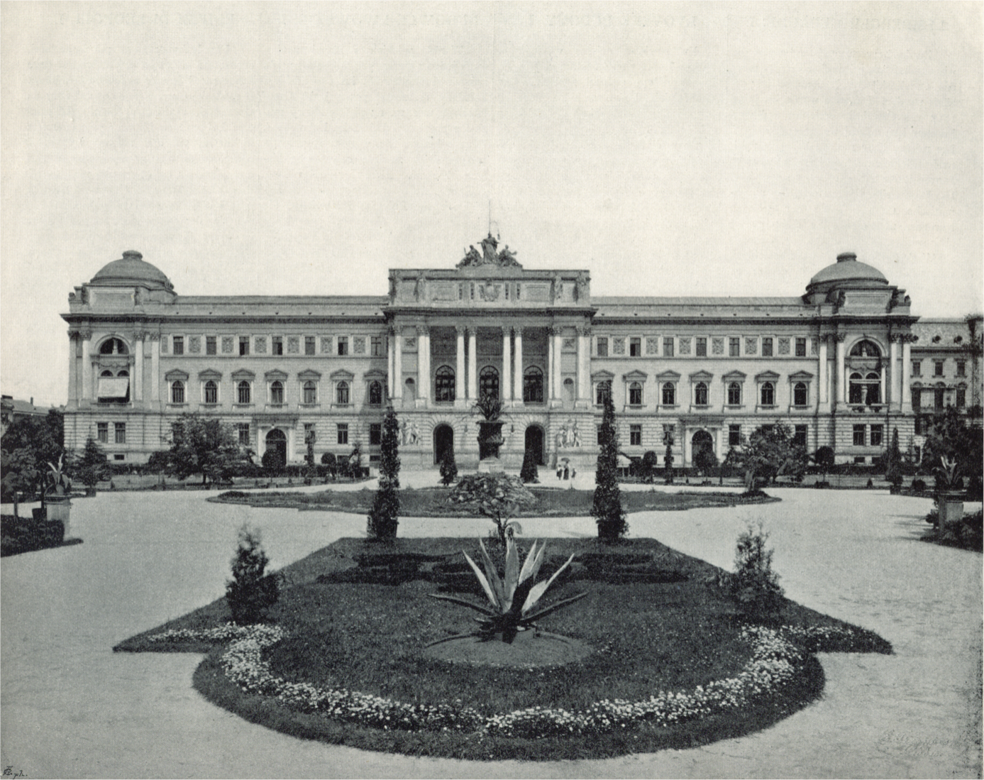 Zdjęcie przedstawia dwupiętrowy, szeroki budynek. Środkowa część składa się z wejścia, do którego prowadzą wysokie kolumny. Przed budynkiem znajduje się plac z ogrodem.