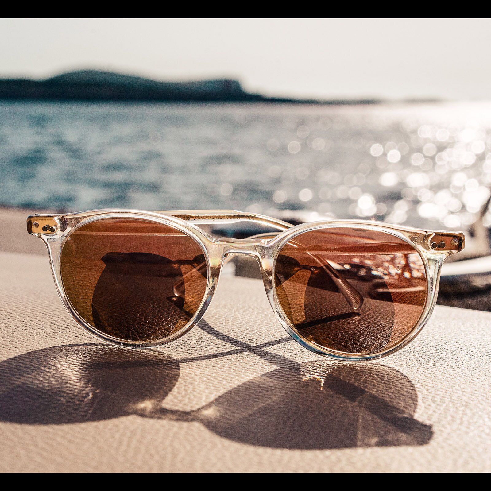 Zdjęcie przedstawia złożone okulary przeciwsłoneczne z przezroczystymi oprawkami, które leżą na kremowej powierzchni etui, a w tle widoczna jest rozmyta, połyskująca w słońcu woda.