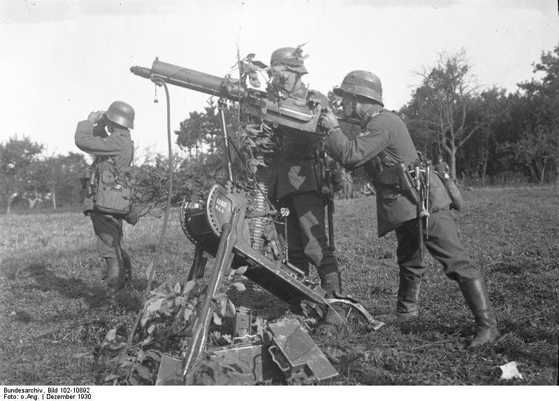 Fotografia przedstawia trzech niemieckich żołnierzy na trawiastym terenie. Pierwszy celuje z wielkiego karabinu na metalowym stojaku, drugi podkłada amunicję. Trzeci stoi trochę dalej i wygląda przez lornetkę.