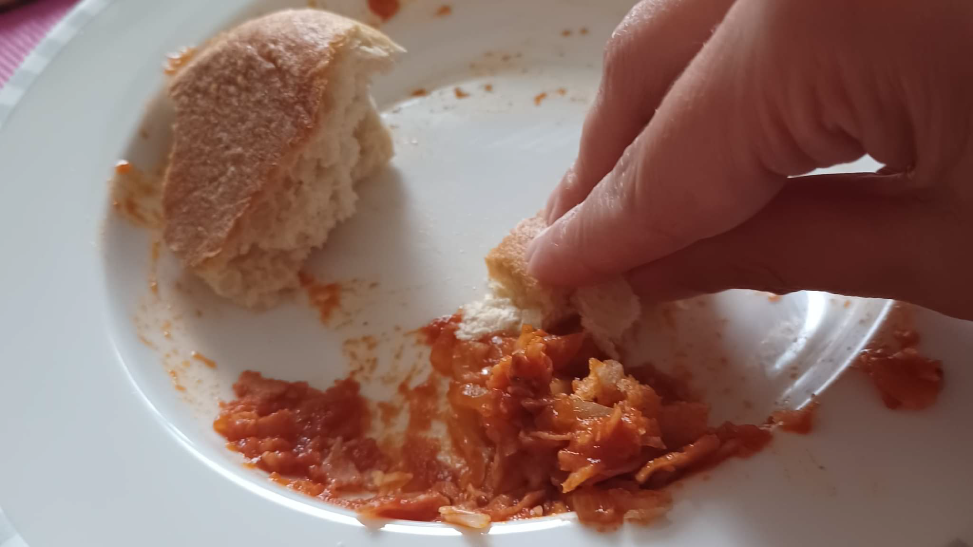 Zdjęcie przedstawia dłoń osoby trzymającą kawałek chleba. Widoczny jest talerz z resztkami jedzenia i sosu. Osoba wybiera tym kawałkiem chleba resztki jedzenia z talerza.