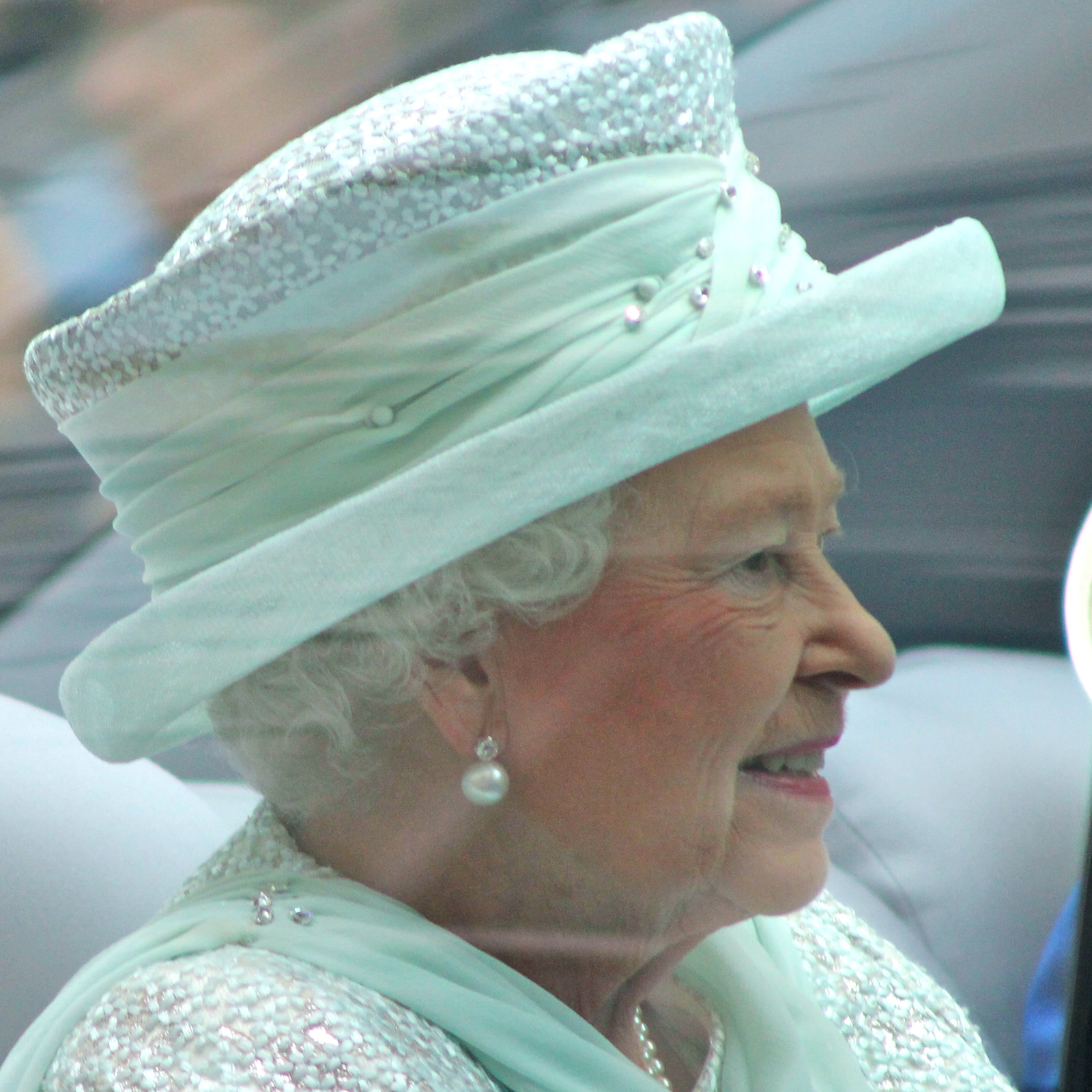 Zdjęcie przedstawia królową Elżbietę II, w ujęciu od ramion w górę. Widoczna jest kobieta w starszym wieku, ubrana w jasnoniebieską górę i jasnoniebieski kapelusz. Zwrócona jest w prawo i patrzy przed siebie. Kobieta ma w uszach kolczyk z perłą i delikatnie się uśmiecha.