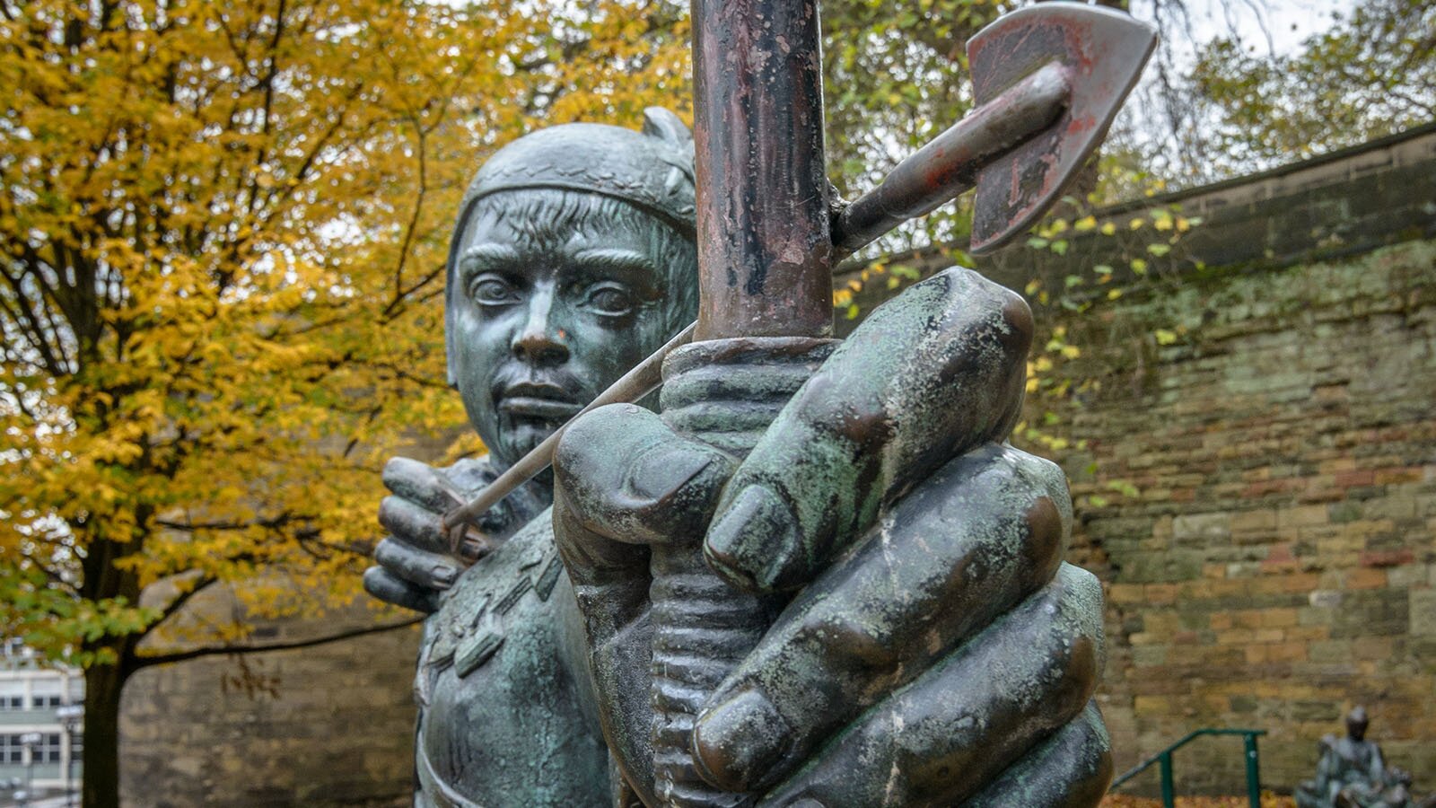 Zdjęcie przedstawia posąg młodego łucznika od klatki piersiowej w górę. W rękach trzyma naprężony łuk ze strzałą, wymierzoną prosto w obiektyw. W tle są drzewa oraz mur z szarej cegły.