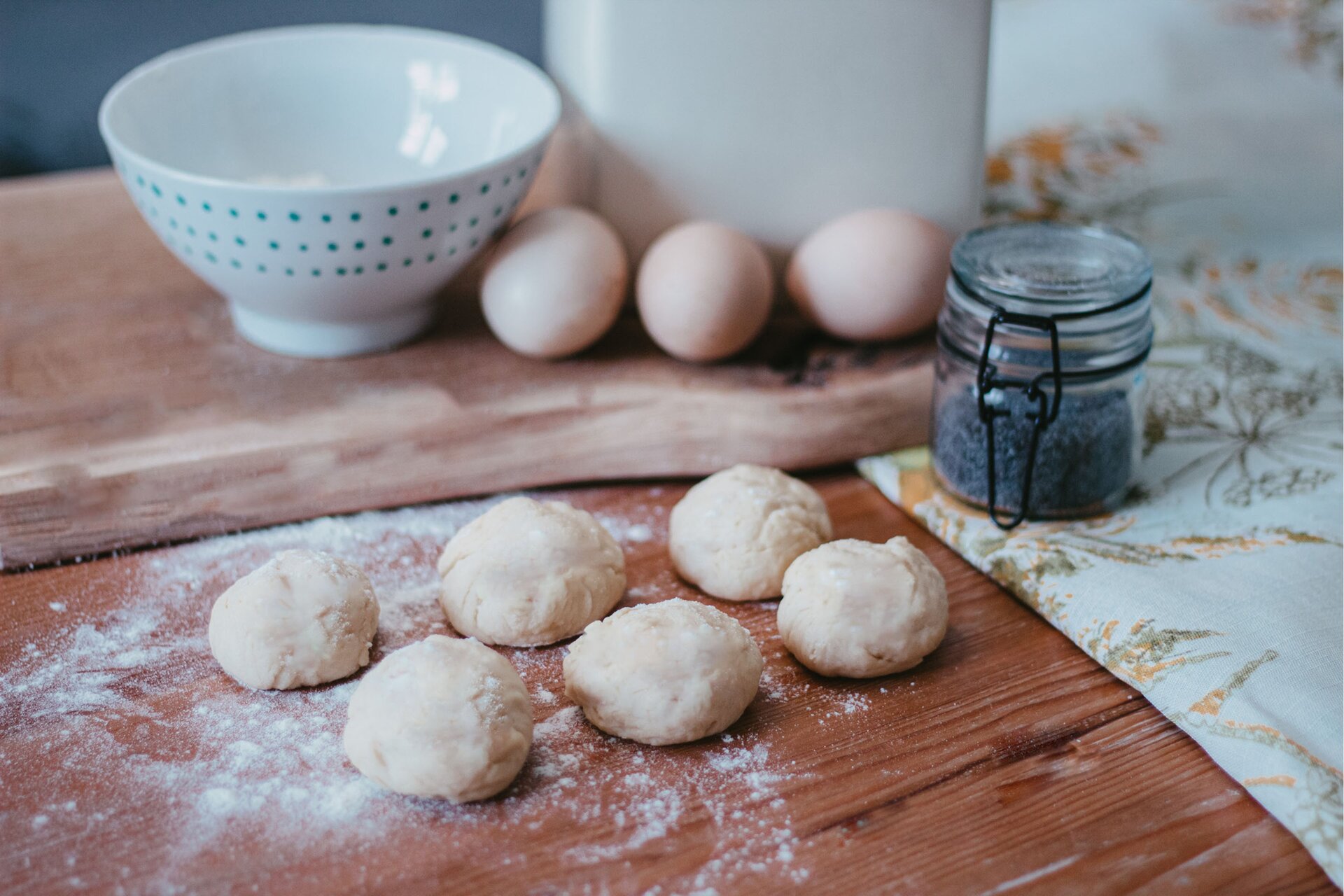 Zdjęcie przedstawia kontynuację kroku drugiego formowania chały kijowskiej. Ujęcie wykonane jest od góry pod skosem. Na ciemnobrązowym drewnianym blacie znajduje się sześć kęsów jasnego ciasta. Mają one kształt małych kulek. Ułożone są poziomo po trzy w górnym i dolnym rzędzie. Kęsy ciasta oprószone sa mąką, która rozsypana jest również dookoła nich na blacie. W prawej części zdjęcia widoczny jest biały obrus w żółte i zielone motywy roślinne na którym stoi szklany przezroczysty słoik wypełniony do połowy niebieskim makiem. W górnej części grafiki znajduje się drewniana jasnobrązowa deska. Stoi na niej biała miska w niebieskie małe kropki. Obok niej po prawej stronie dostrzec można trzy jajka. Za nimi znajduje się biały wysoki prostokątny pojemnik.