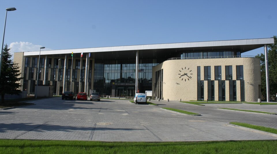 Kliknij żeby powiększyć Zdjęcie, które przedstawia siedzibę Urzędu Gminy Michałowice w Regułach. Nowy, duży budynek. W znacznej części przeszklony. Po prawej stronie znajduje się ściana. Na ścianie budynku widoczny zegar i przeszklone okna. Przed wejściem do budynku znajduje się parking. Na parkingu są zaparkowane auta. Przed parkingiem znajduje się trawnik.