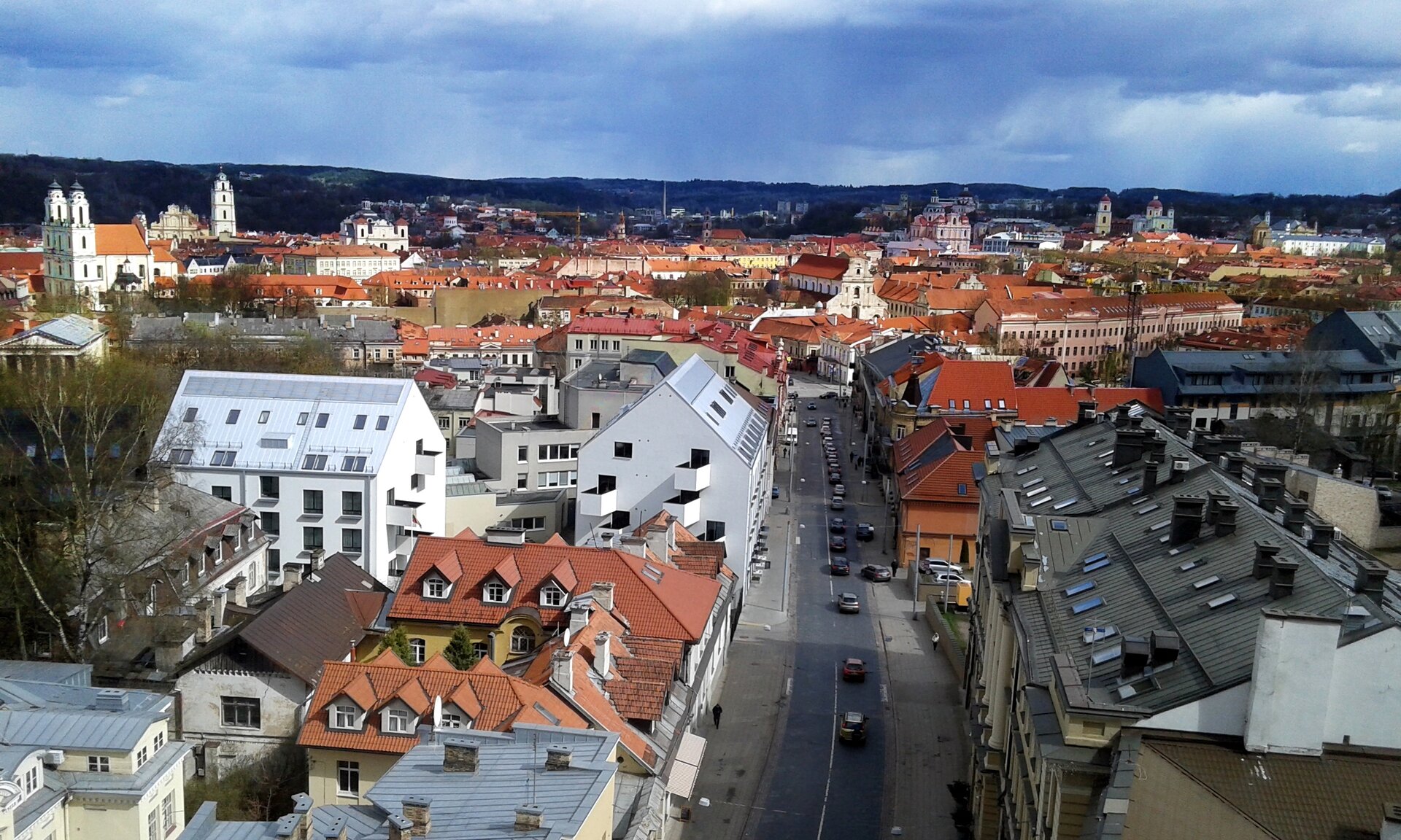 Na zdjęciu panorama miasta. Nad budynkami górują wieżyczki kościołów. Po środku fotografii między budynkami ulica. W tle teren zalesiony.