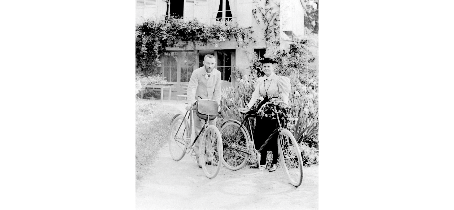 Na czarnobiałym zdjęciu widoczni mężczyzna oraz kobieta, pozujący do zdjęcia z rowerami. W tle budynek otoczony roślinnością.