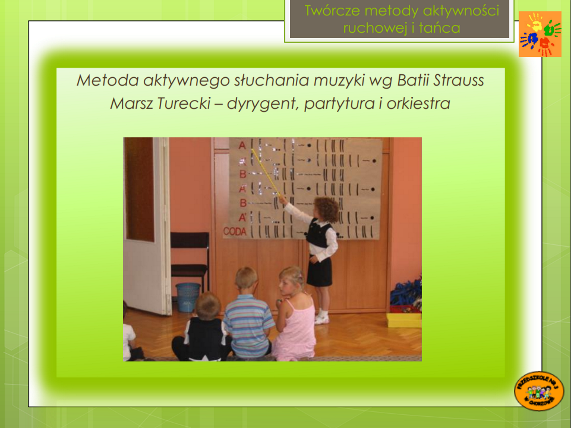 Ilustracja przedstawia zdjęcie, które znajduje się w zielonej ramce i podpisane jest u góry: "Twórcze metody aktywności ruchowej i tańca. Metoda aktywnego słuchania muzyki według Batii Strauss Marsz Turecki - dyrygent, partytura i orkiestra". Na zdjęciu stoi mała dziewczynka, która pokazuje wskaźnikiem na partyturę wykonaną na dużym arkuszu papieru szarego. Partytura składa się z przyklejonych liter, linii poziomych i pionowych oraz kropek. Arkusz papieru wisi na tle drewnianej szafy. Trójka dzieci siedzi na drewnianej podłodze przed stojącą dziewczynką.