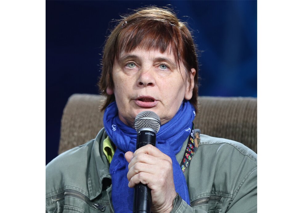 Fotografia barwna przedstawiająca Janinę Ochojską. Ma na szyi fioletowawy szal, w dłoni trzyma mikrofon, do którego coś mówi. Siedzi na fotelu, ubrana jest w zielonkawą kurtkę.