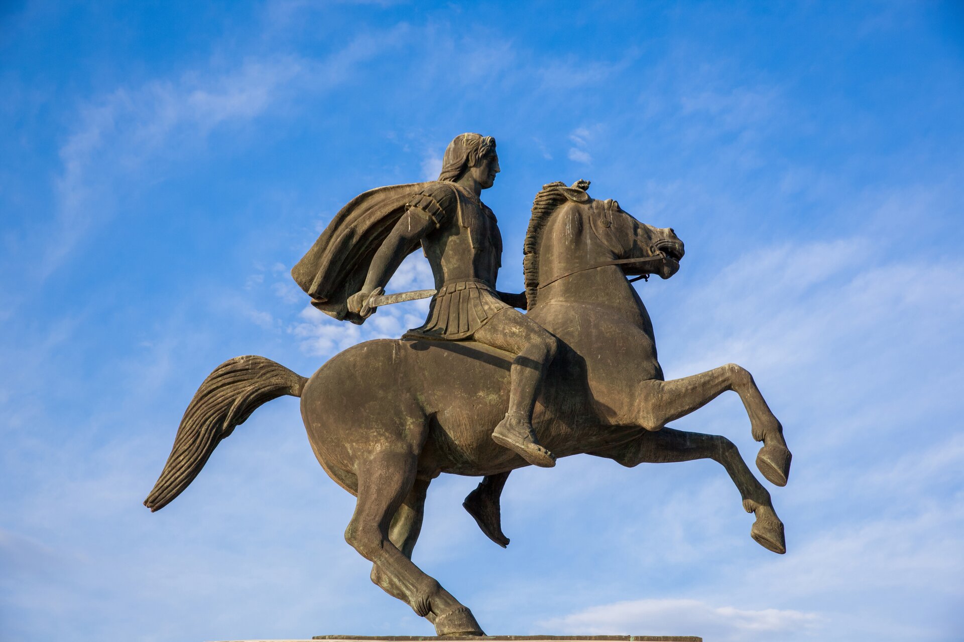 Ilustracja przedstawia pomnik Aleksandra Wielkiego na koniu Bucefale. Koń stoi na dwóch nogach, Aleksander Wielki jedną ręką trzyma wodze, w drugiej trzyma miecz.