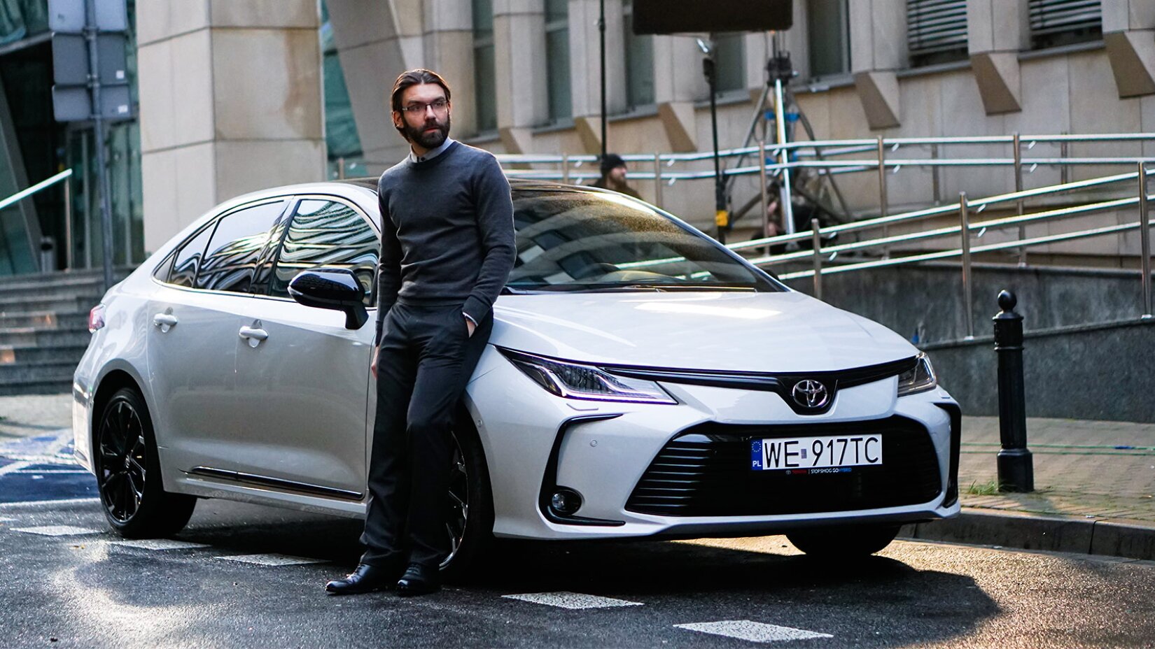 Kadr z reklamy Toyoty Corolli GR Sport, na którym widać mężczyznę w średnim wieku opierającego się o białe auto z czarnymi detalami. Mężczyzna opiera się o maskę auta tuż przy przednich drzwiach pojazdu. Ma na sobie czarne eleganckie spodnie, garniturowe buty, a także szary sweter, spod którego wystaje kołnierz jasnoniebieskiej koszuli. Jego lewa ręka spoczywa w kieszeni spodni, mężczyzna patrzy w bok. Jego włosy zaczesane są do tyłu, z przedziałkiem pośrodku, a jego zarost jest średniej długości. Można dostrzec okulary w cienkiej oprawie. W tle widać budynek oraz prowadzące do niego schody, a także statyw. 