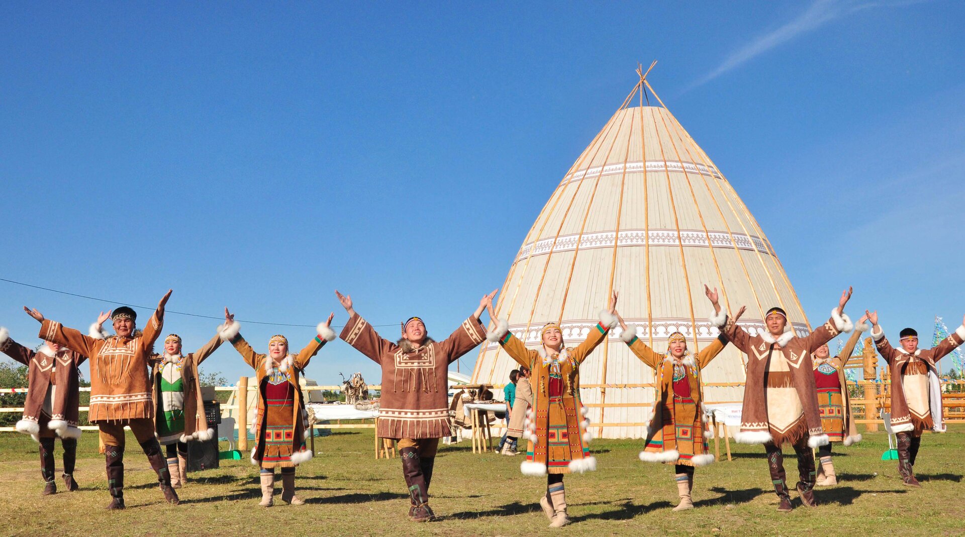 Na zdjęciu grupa osób ubranych w brązowe kubraki ozdobione frędzlami. Wszyscy mają dłonie uniesione w górę. 