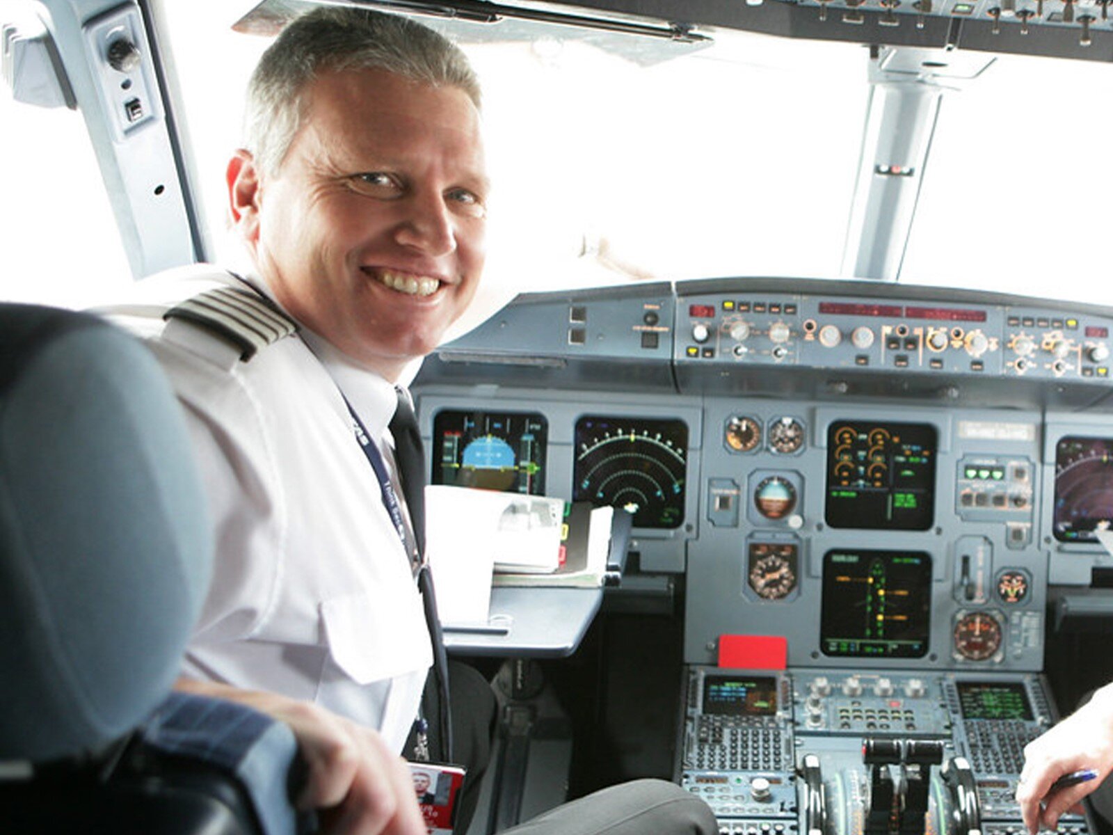 Zdjęcie przedstawia pilota w średnim wieku, siedzącego w kokpicie samolotu. Mężczyzna jest odwrócony w stronę obiektywu i szeroko się uśmiecha. Jest ubrany w białą koszulę z czarnym krawatem, będącą częścią munduru pilota. Na ramieniu widoczny jest czarny pagon z czterema białymi paski. Przed nim widoczna jest aparatura sterowania samolotu z tarczami z zegarami, wyświetlaczami, przyciskami oraz dźwigniami.