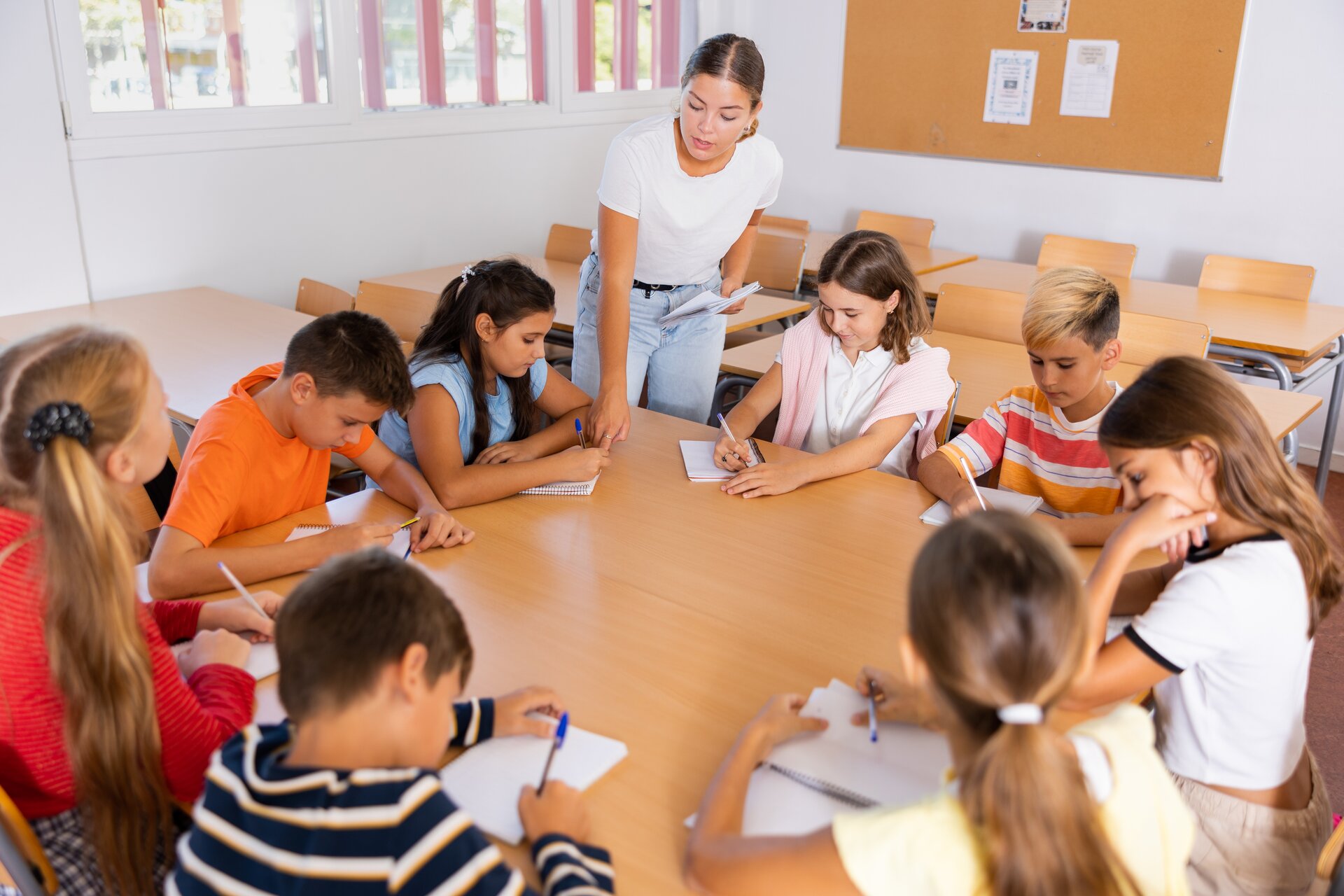 Fotografia przedstawia grupę uczniów siedzących w kole. Uczniowie piszą notatki, a nauczycielka kontroluje ich pracę. Nauczycielka to młoda kobieta ubrana w białą koszulkę oraz jasne długie spodnie jeansowe.